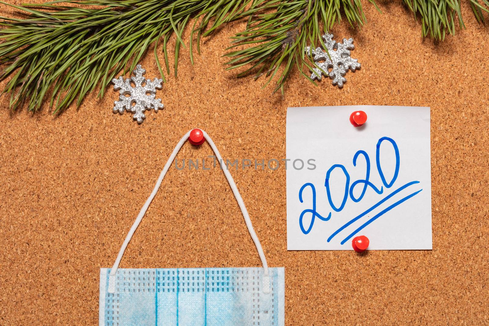 Medical mask and white sticky note with 2020 pinned on notice board which is decorated with pine twigs and snowflakes. Healthcare, christmas, new years celebration season concept. New reality concept by DamantisZ