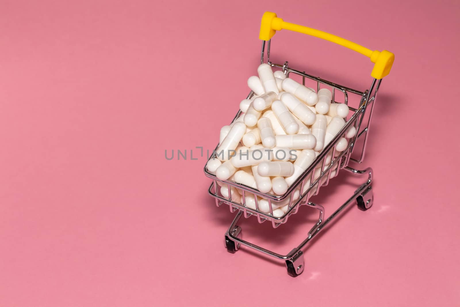 High angle shot of a small shopping cart full of white pills. Pink background, copy space. Shopping online, buying medicine, pharmaceutical business concepts.