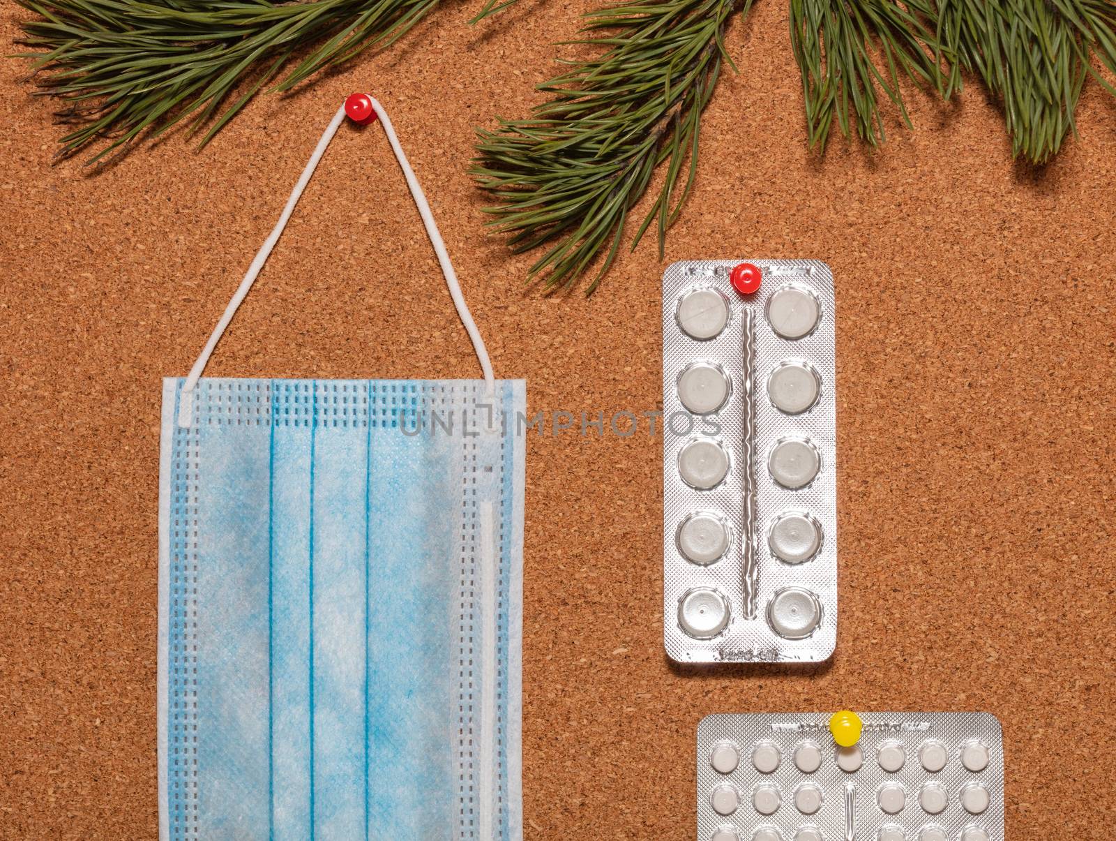 Medical mask and white pills pinned on a corkwood notice board which is decorated with pine twigs at the top. Healthcare, christmas, new years celebration, new normal concepts.