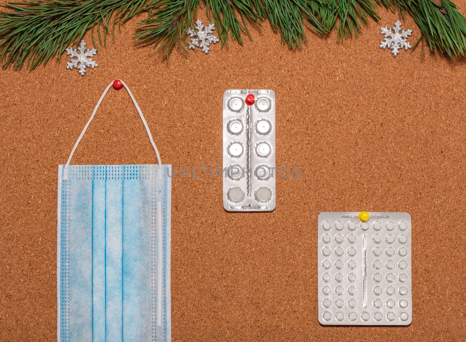 Medical mask and white pills pinned on a corkwood notice board which is decorated with pine twigs and silver snowflakes at the top. Healthcare, christmas, new years celebration, new normal concepts.