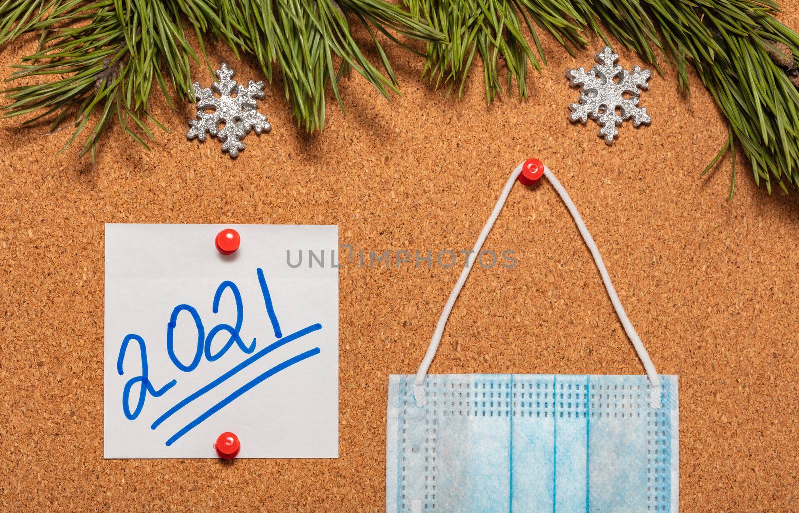 Medical mask and white sticky note with 2021 pinned on notice board which is decorated with pine twigs and snowflakes. Healthcare, christmas, new years celebration season concept. New reality concept