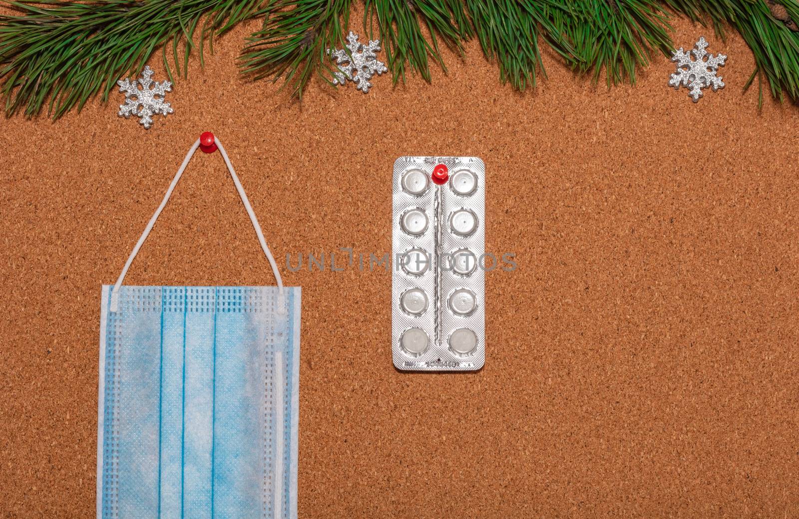 Medical mask and white pills pinned on a corkwood notice board which is decorated with pine twigs and silver snowflakes at the top. Healthcare, christmas, new years celebration, new normal concepts.