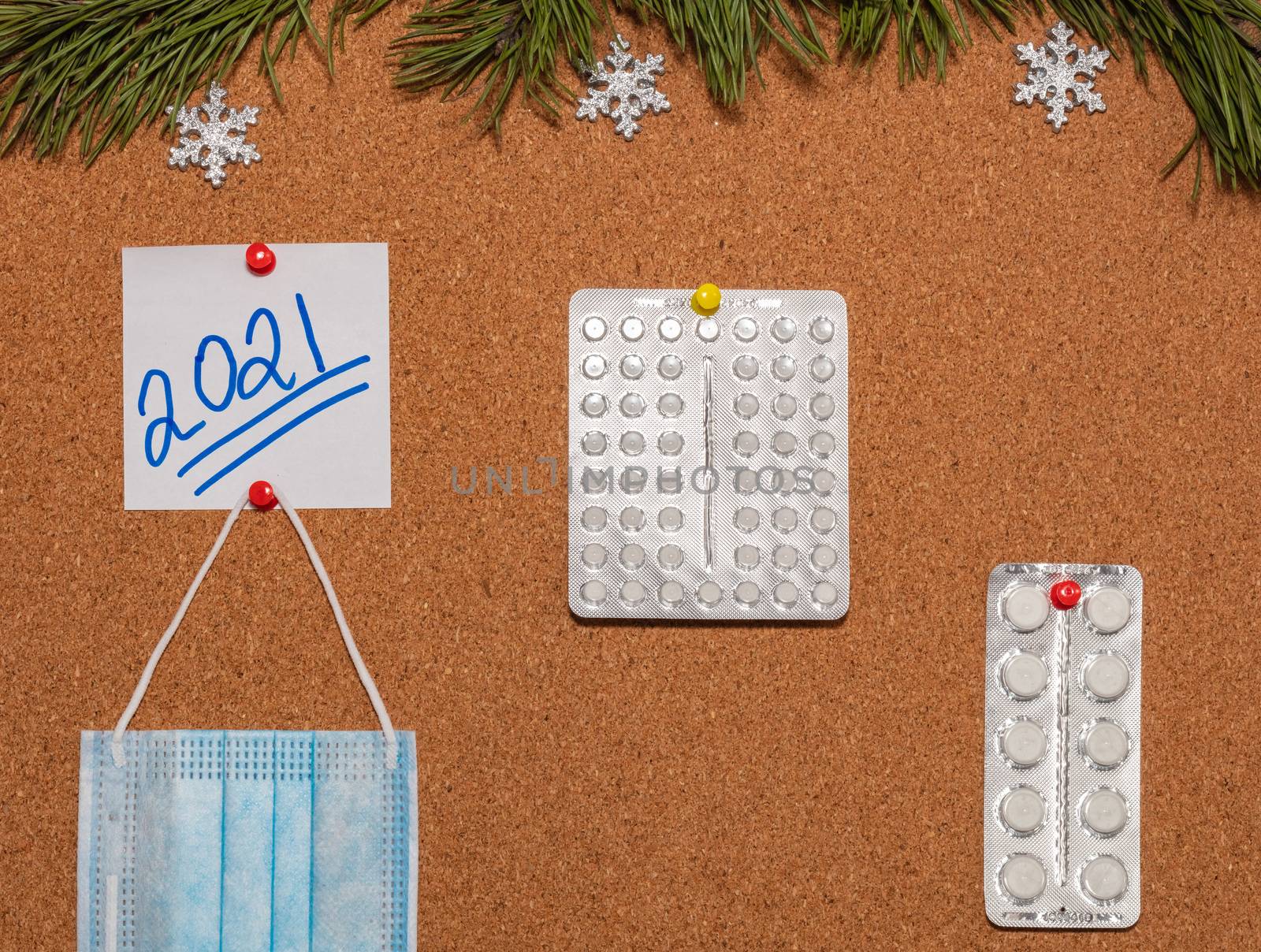 Medical mask, white pills, white sticky note with 2021 pinned on notice board which is decorated with pine twigs and snowflakes. Healthcare, christmas, new years celebration, new normal concepts.