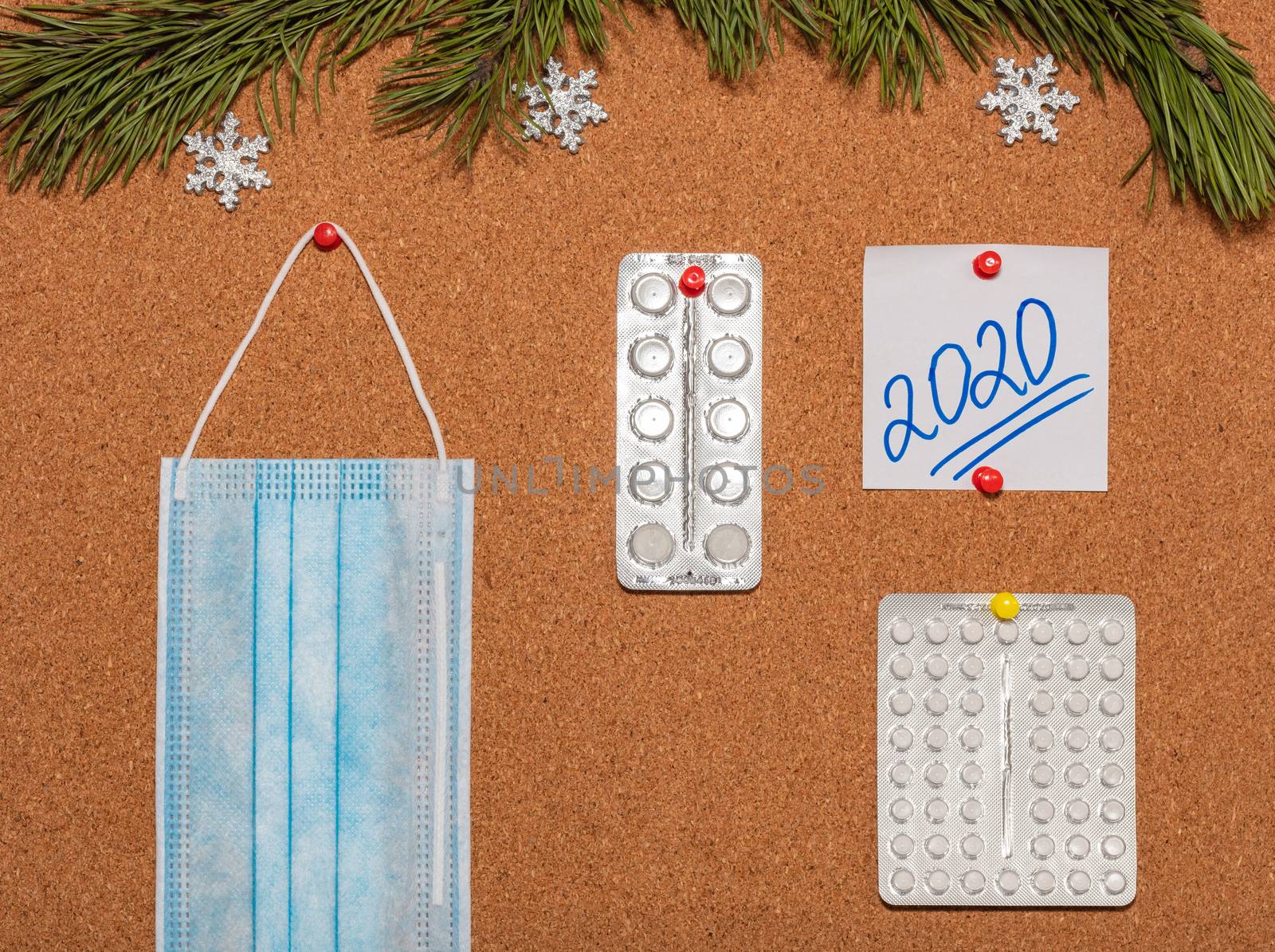 Medical mask, white pills, white sticky note with 2020 pinned on notice board which is decorated with pine twigs and snowflakes. Healthcare, christmas, new years celebration, new normal concepts.