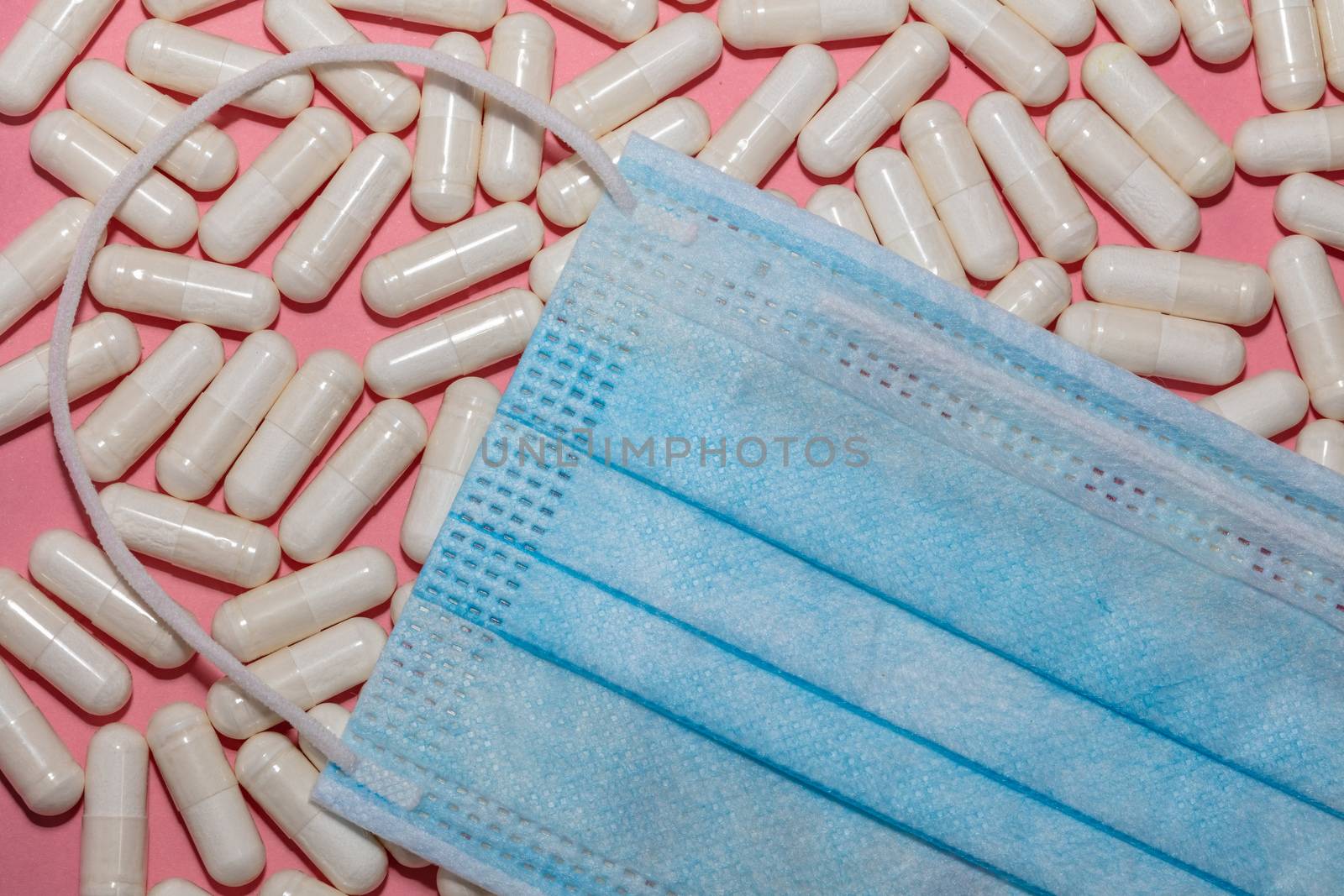 Top close up shot of white pills and blue medical mask on top of them on pink background. Healthcare, medical and pharmaceutical concept. New normal and reality concept.