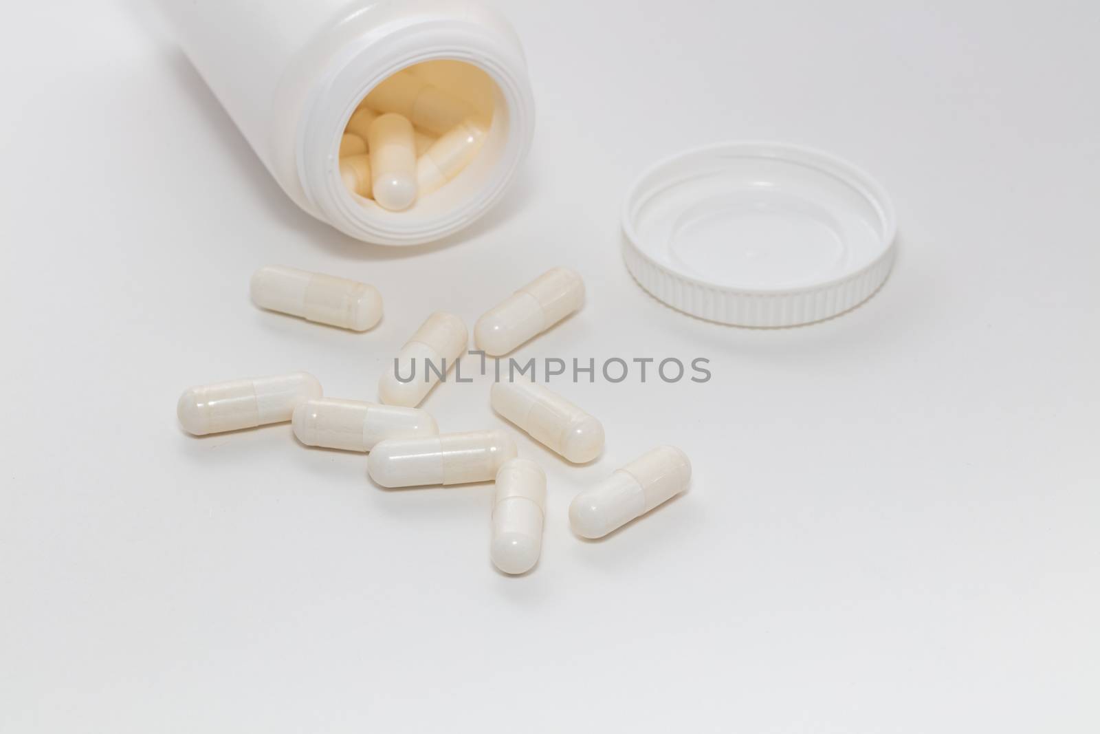 Bunch of white scattered pills on white background. White pills container and cap next to them slightly out of focus in the background. Pharmaceutical business and medicine sale concepts. by DamantisZ
