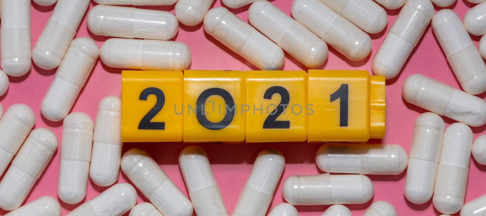 Top close up shot of white pills surrounding yellow cubes with black digits which form 2021 year. Pink background. Healthcare and pharmaceutical concept. New normal and reality concept. Banner size. by DamantisZ