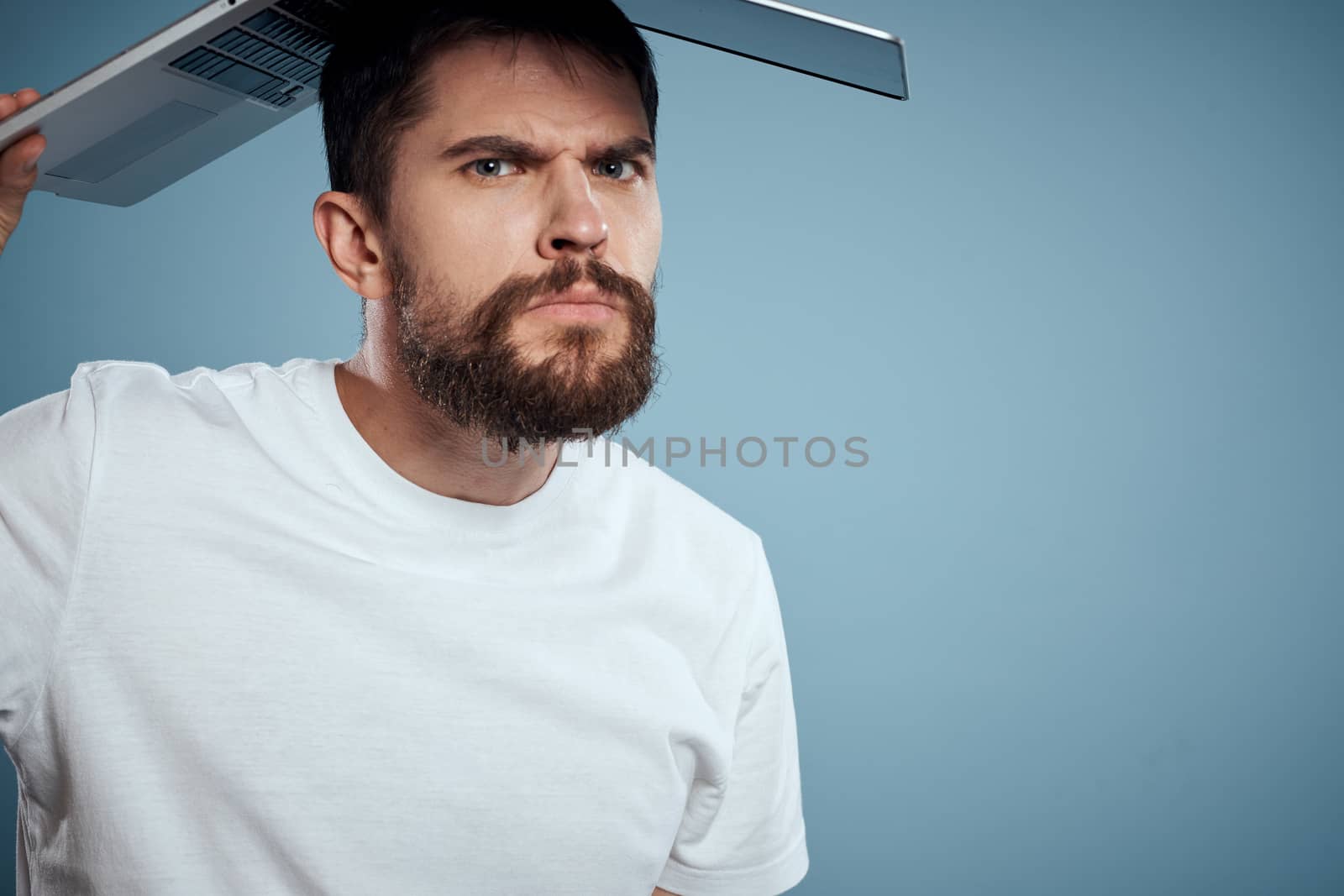 Emotional man with an open laptop above his head On a blue background in a white T-shirt, cropped view by SHOTPRIME