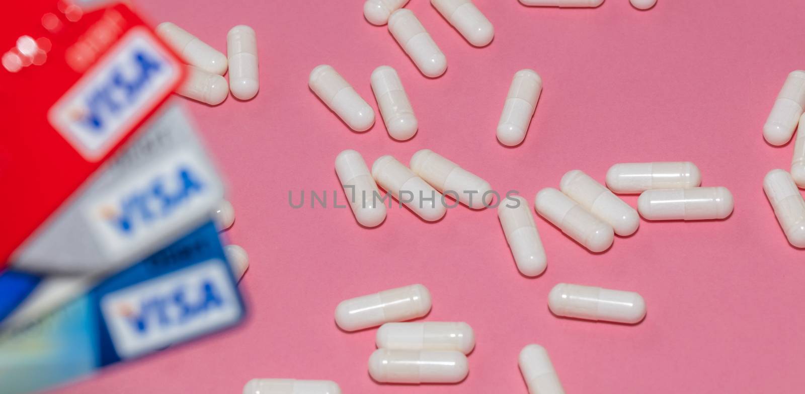 Barnaul, Russia - October 13, 2020: High angle shot of white pills on pink background with three red, silver, blue visa debit cards out of focus in the foreground. Pharmaceutical business concept.
