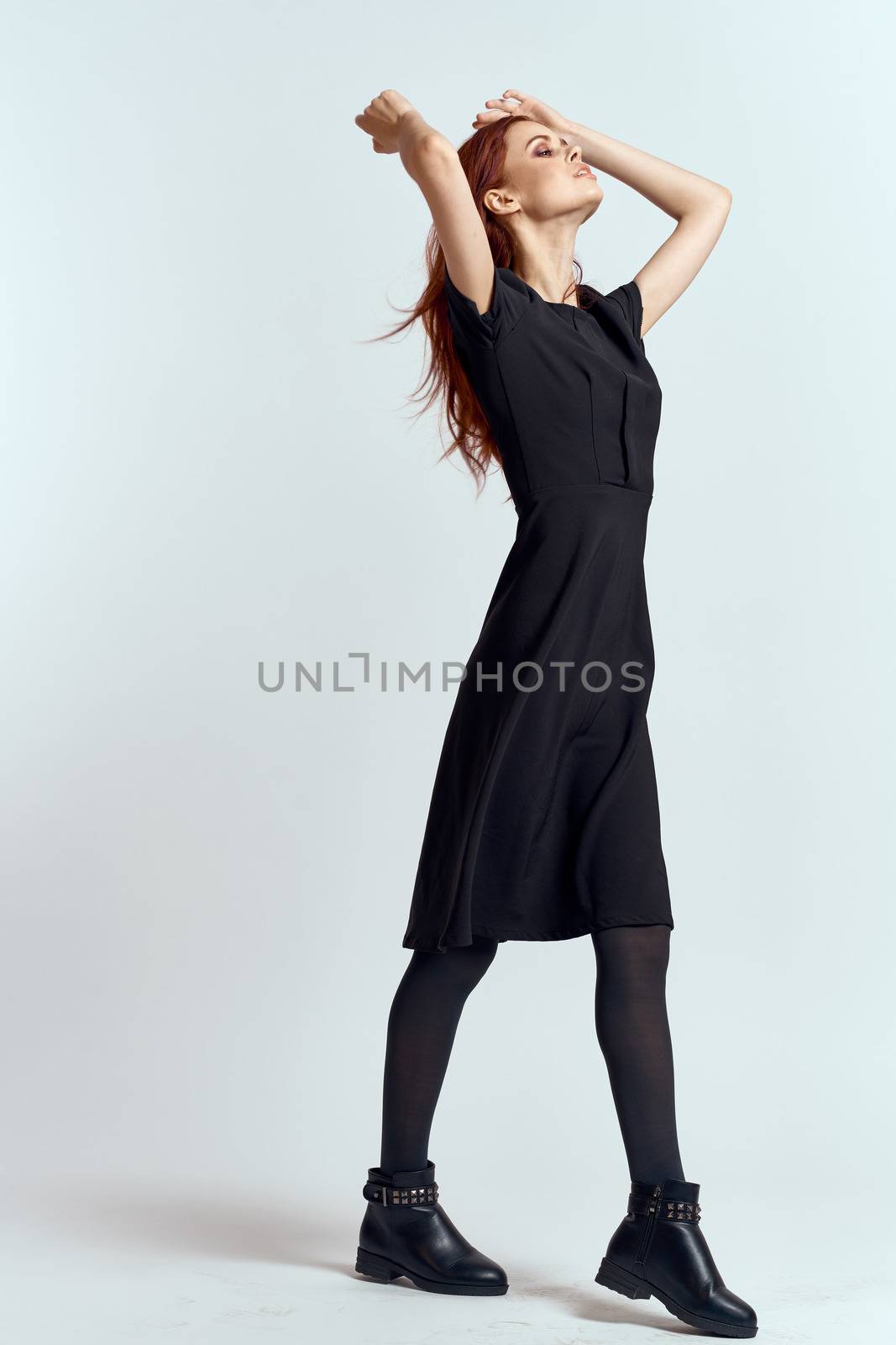 A woman in a black dress on a light background and pantyhose shoes red hair and pose in full growth by SHOTPRIME