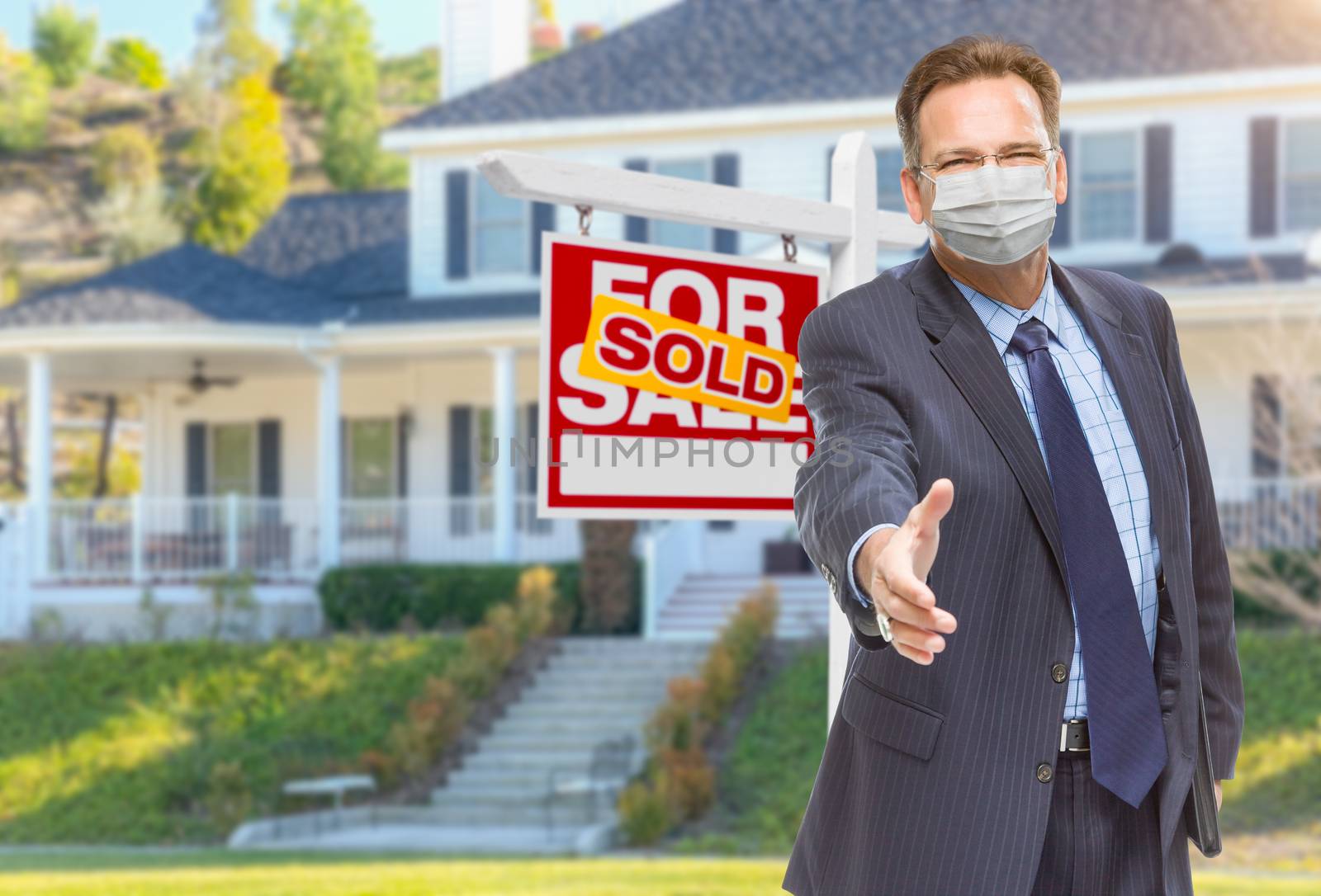 Male Real Estate Agent Reaching for Hand Shake Wearing Medical Face Mask with Sold For Sale Sign Behind.