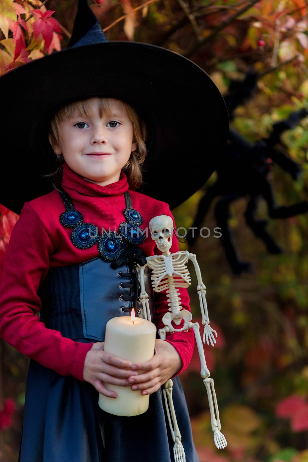 Little girl in a witch costume holding a skeleton on a halloween party in the garden