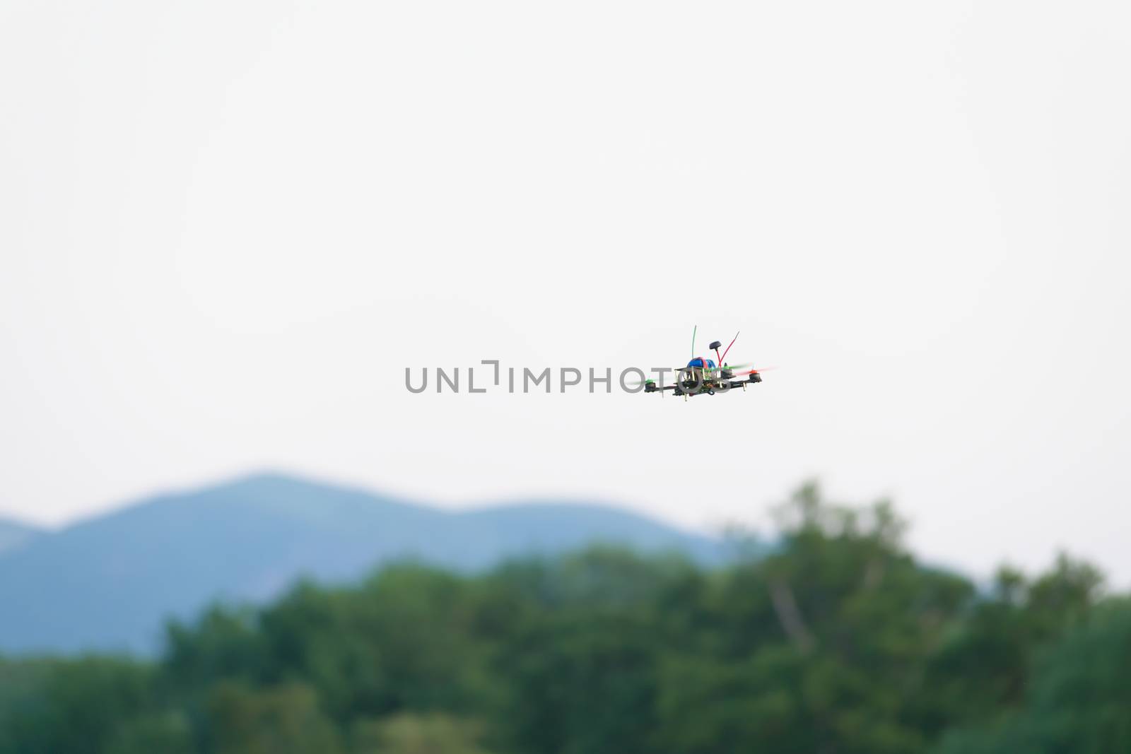 Colorful propellers lift this racing FPV quad copter drone through the air. Plain white sky background with blurred hills and trees beneath