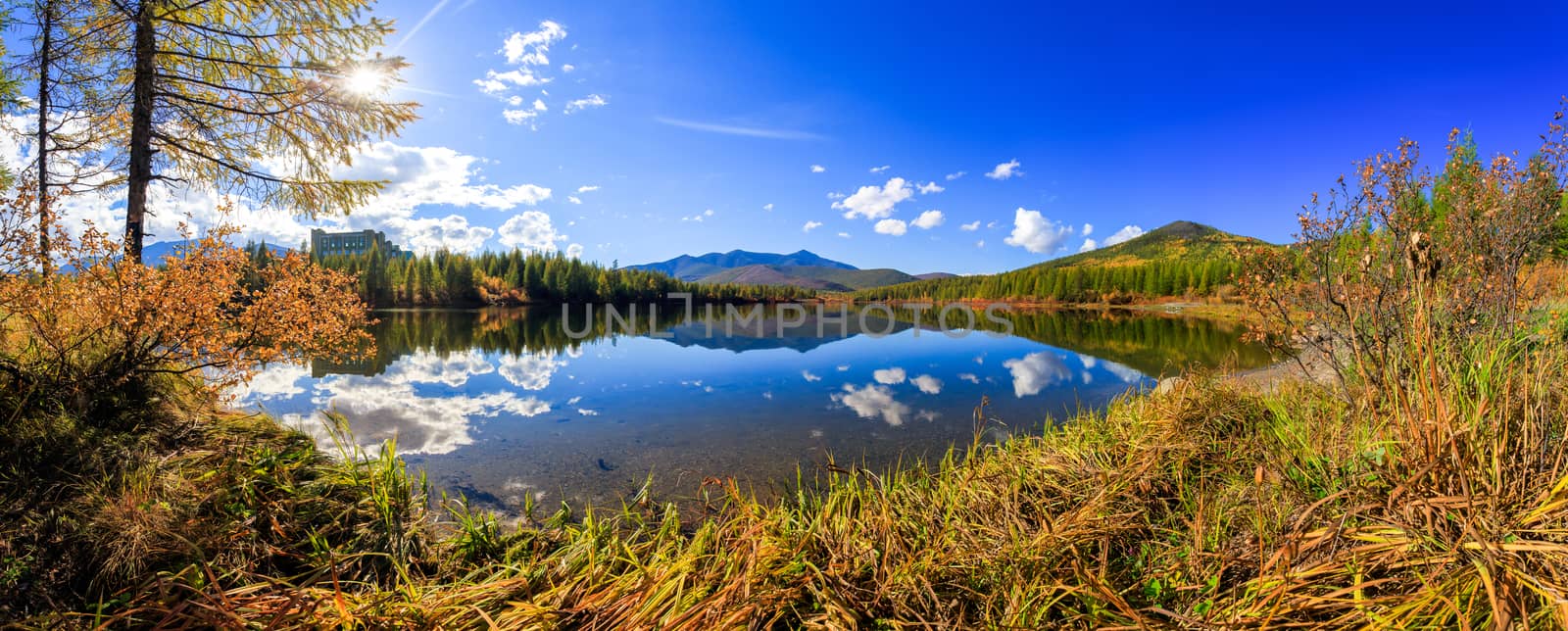 The nature of the Magadan region. Panoramic shot. A beautiful flat surface of the lake against the background of colored hills and the blue sky. Fascinating view of the forest lake