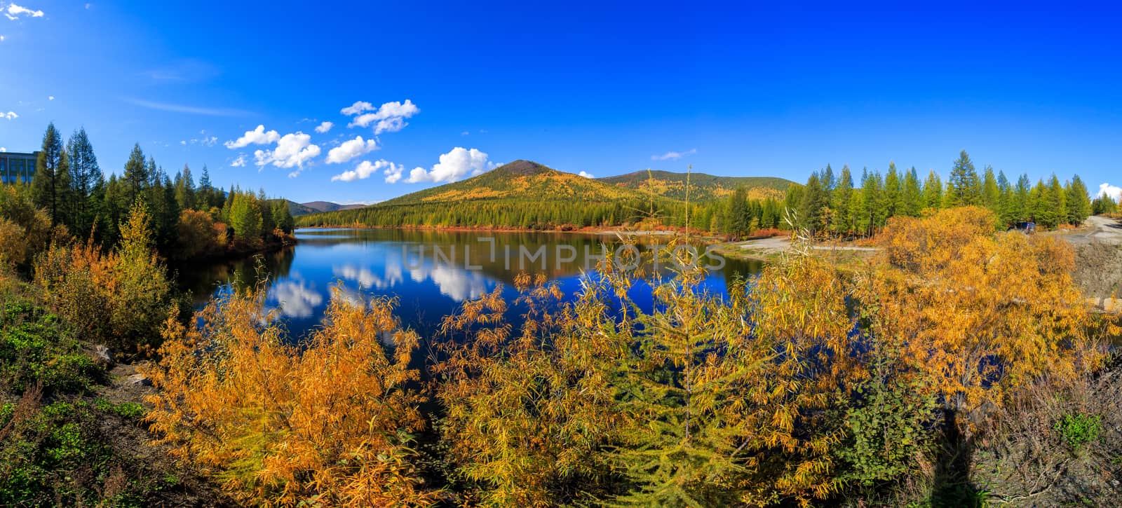 The nature of the Magadan region. Panoramic shot. A beautiful flat surface of the lake against the background of colored hills and the blue sky. Fascinating view of the forest lake