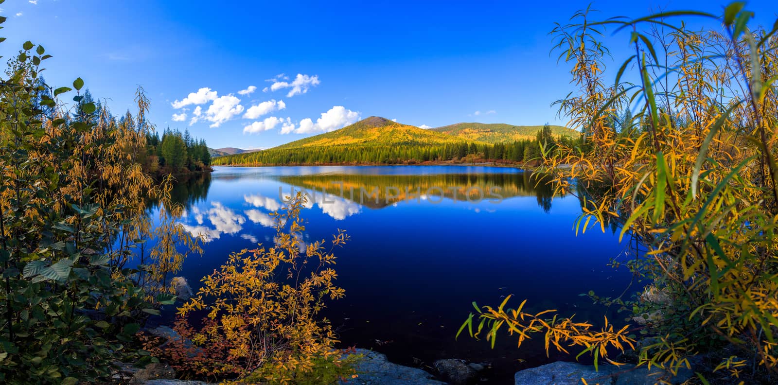 Panoramic shot. A beautiful flat surface of the lake against the background of colored hills and the blue sky. Fascinating view of the forest lake by PrimDiscovery