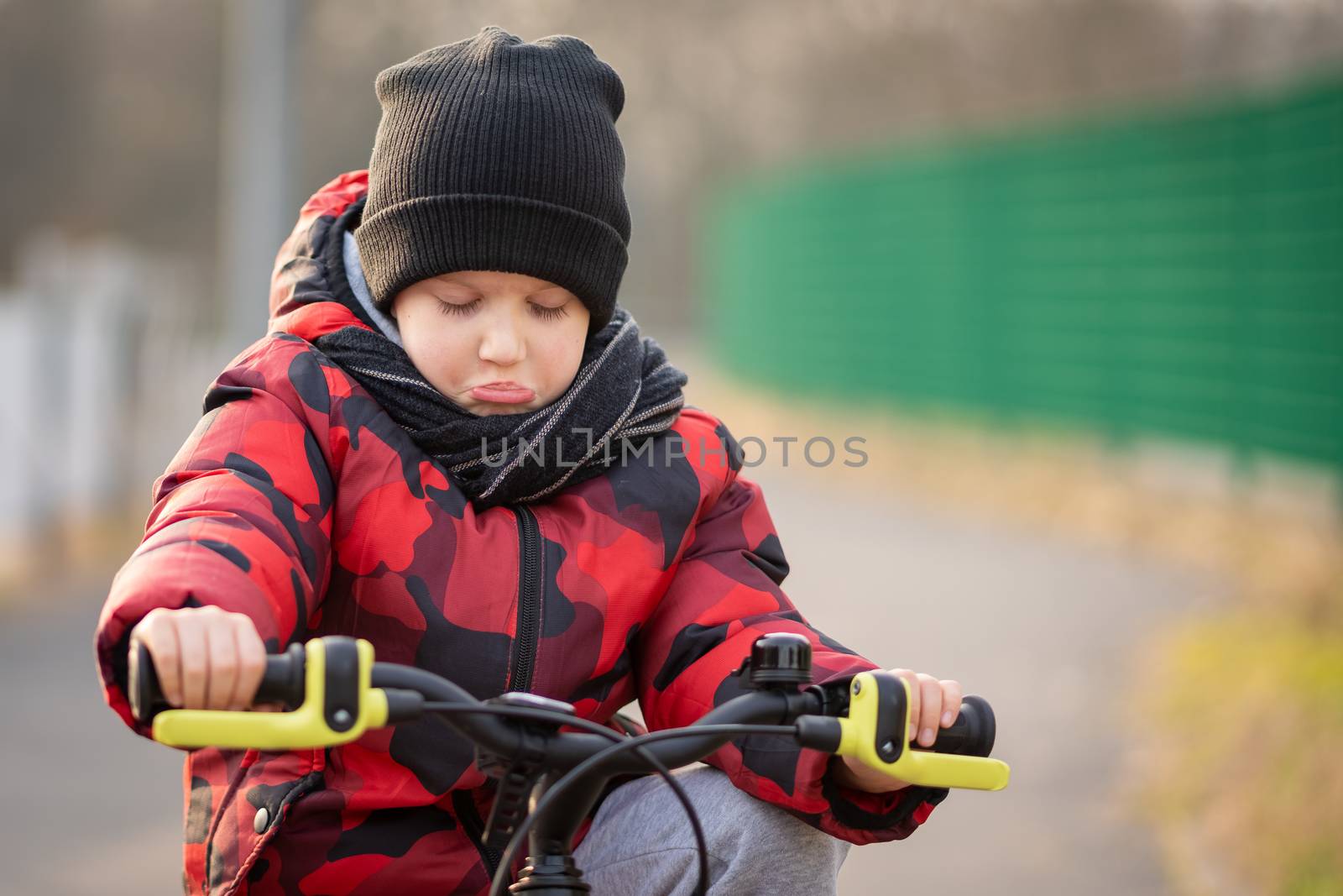 Sad, angry boy with hat and scarf sitting on bicycle at park.
