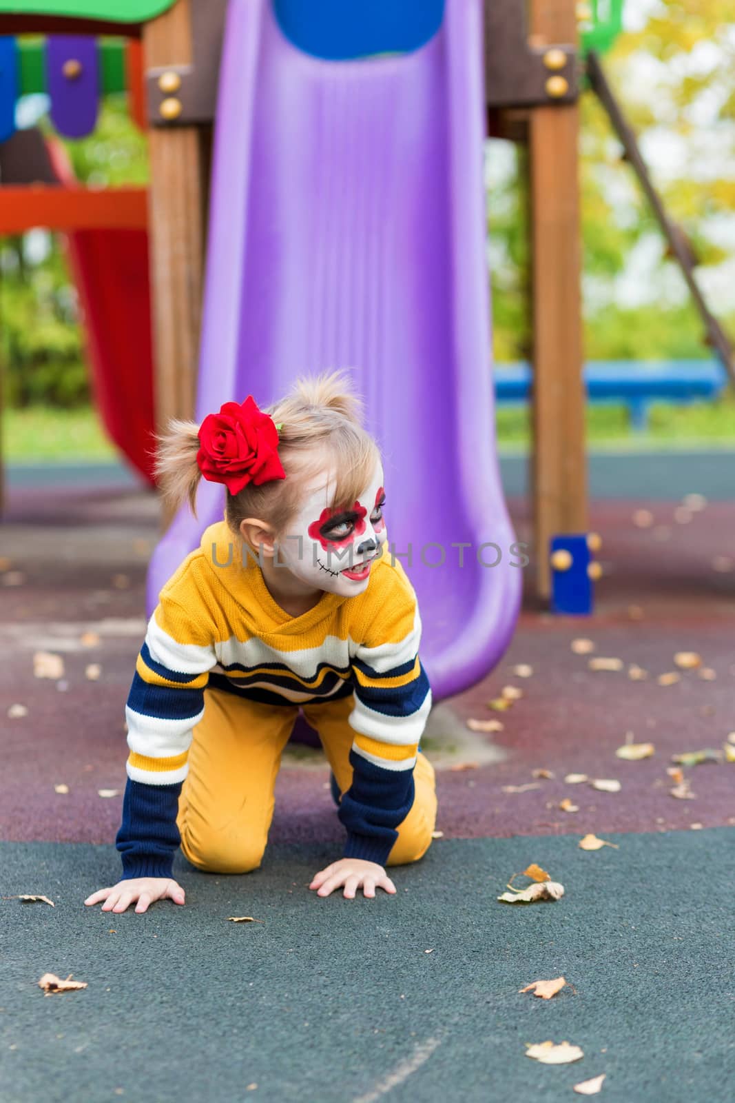 A little preschool girl with Painted Face, rides a slide on the playground, celebrates Halloween or Mexican Day of the Dead.