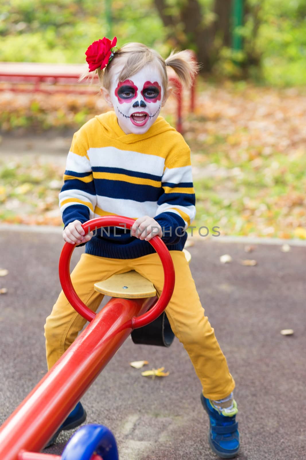 A little preschool girl with Painted Face, rides on a swing in the playground, celebrates Halloween or Mexican Day of the Dead.