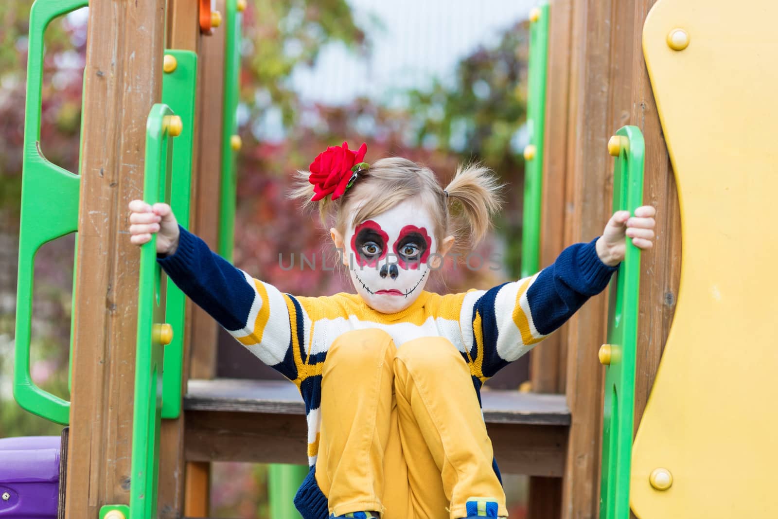 A little preschool girl with Painted Face, shows funny faces on the playground, celebrates Halloween or Mexican Day of the Dead.