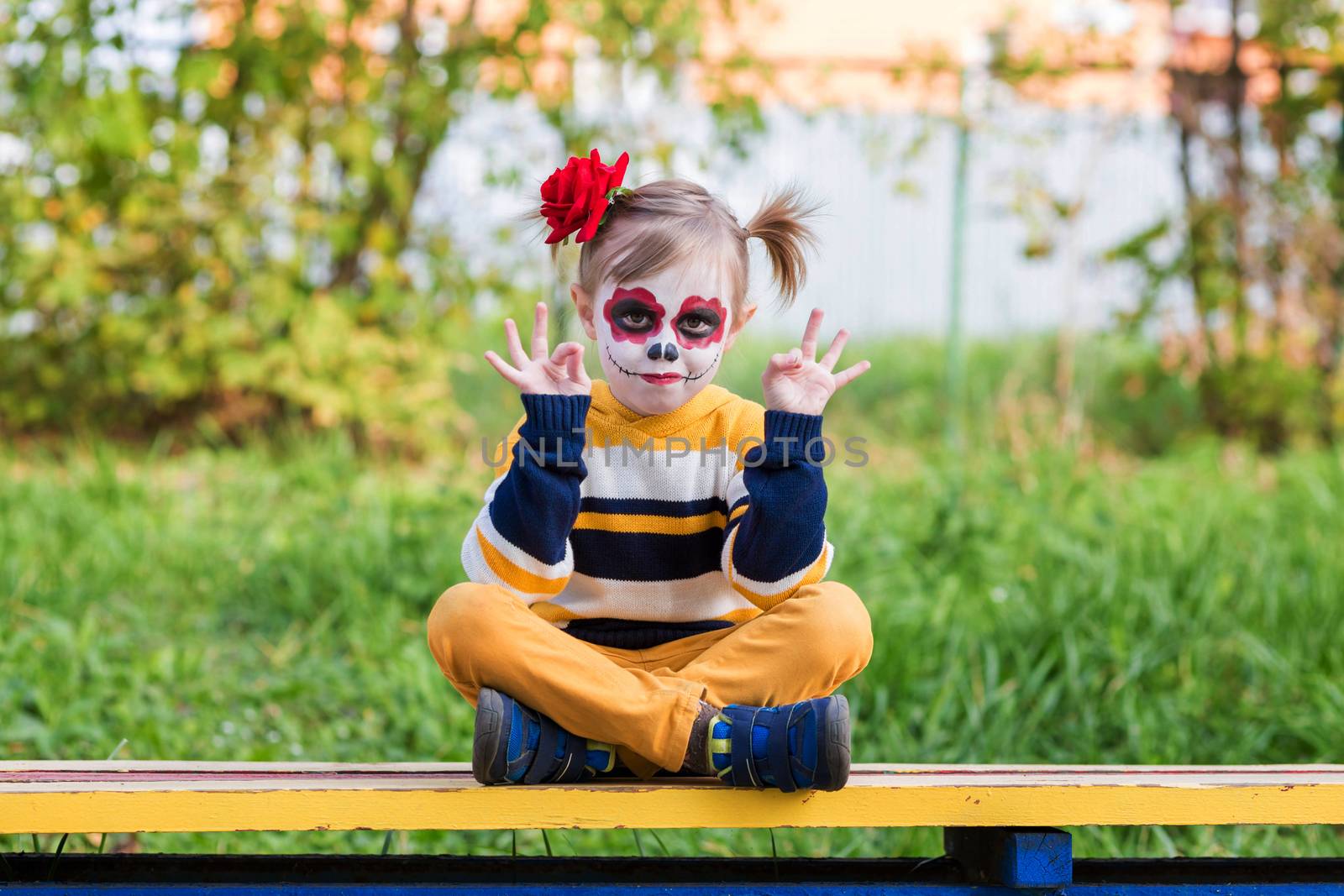 A little preschool girl with Painted Face, sitting on a bench in lotus position on the playground, celebrates Halloween or Mexican Day of the Dead.