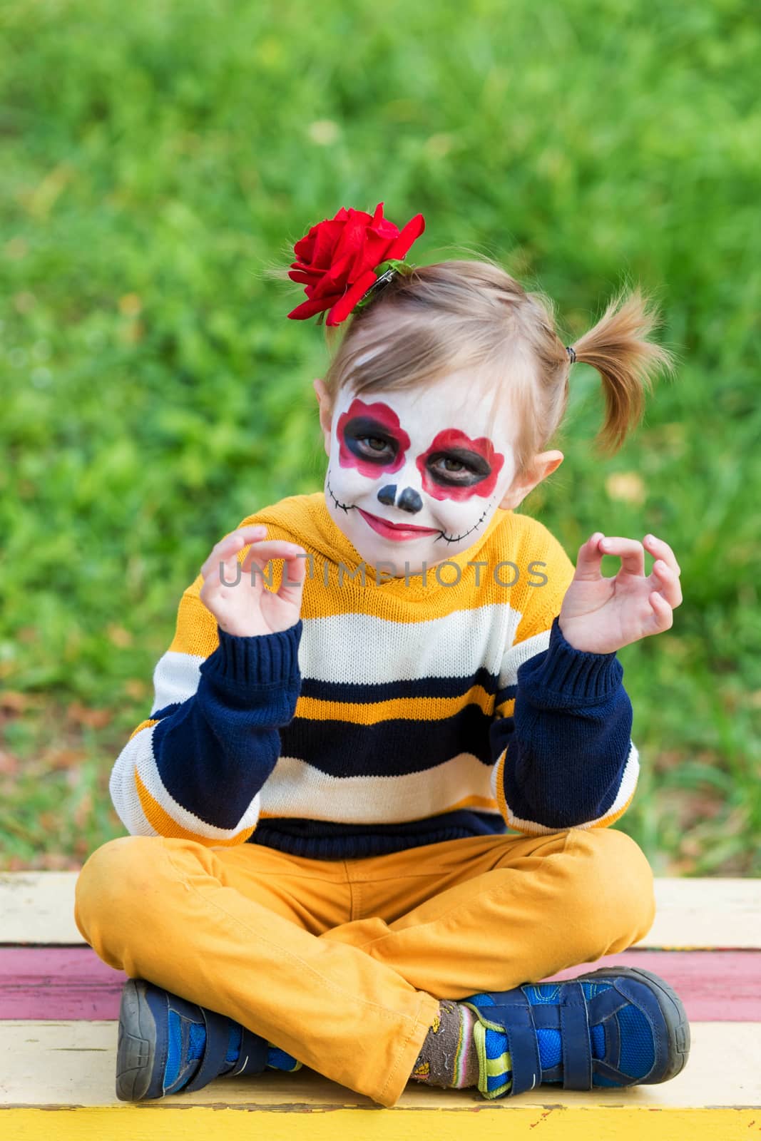 A little preschool girl with Painted Face, sitting on a bench in lotus position on the playground, celebrates Halloween or Mexican Day of the Dead.