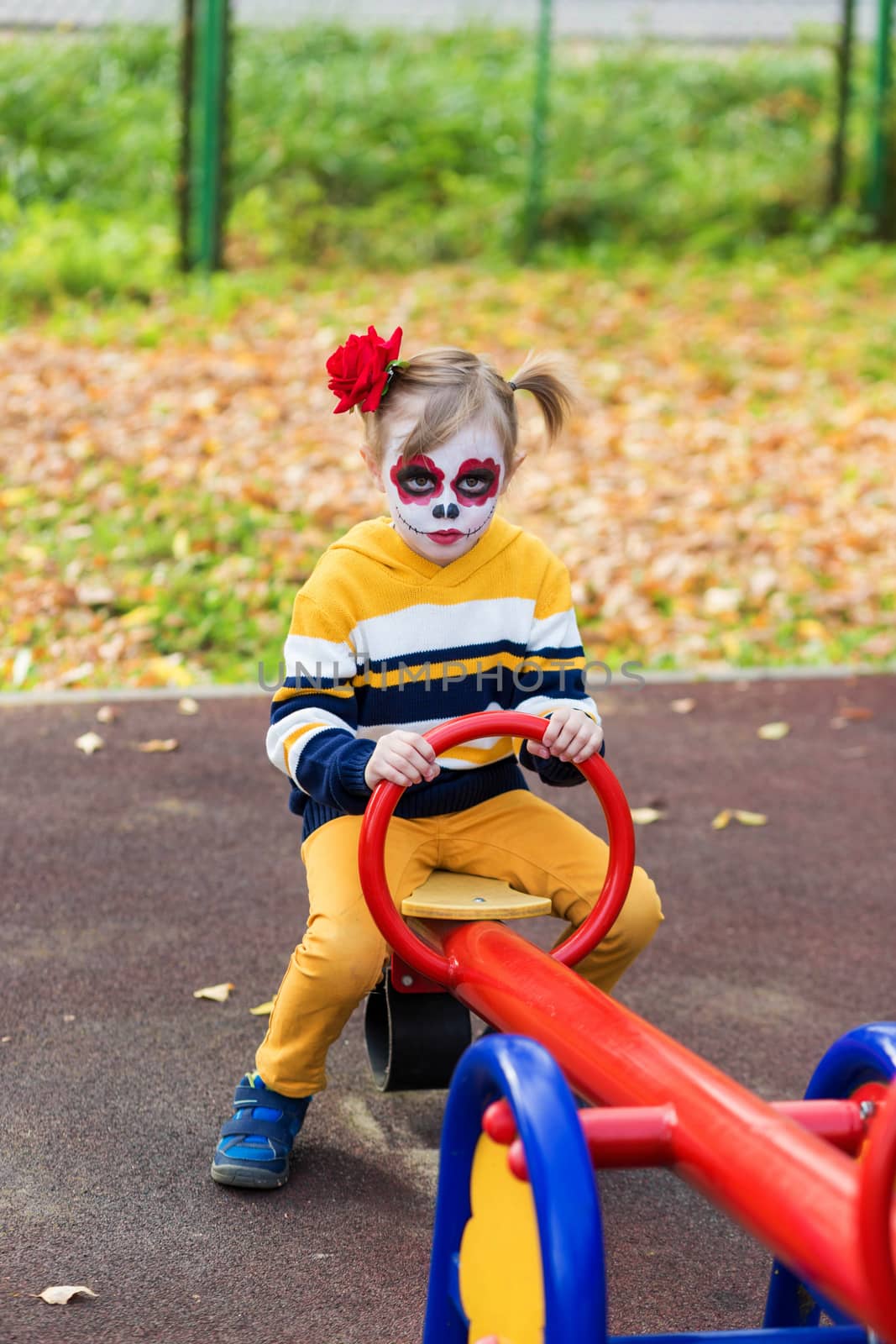 A little preschool girl with Painted Face, rides on a swing in the playground, celebrates Halloween or Mexican Day of the Dead.