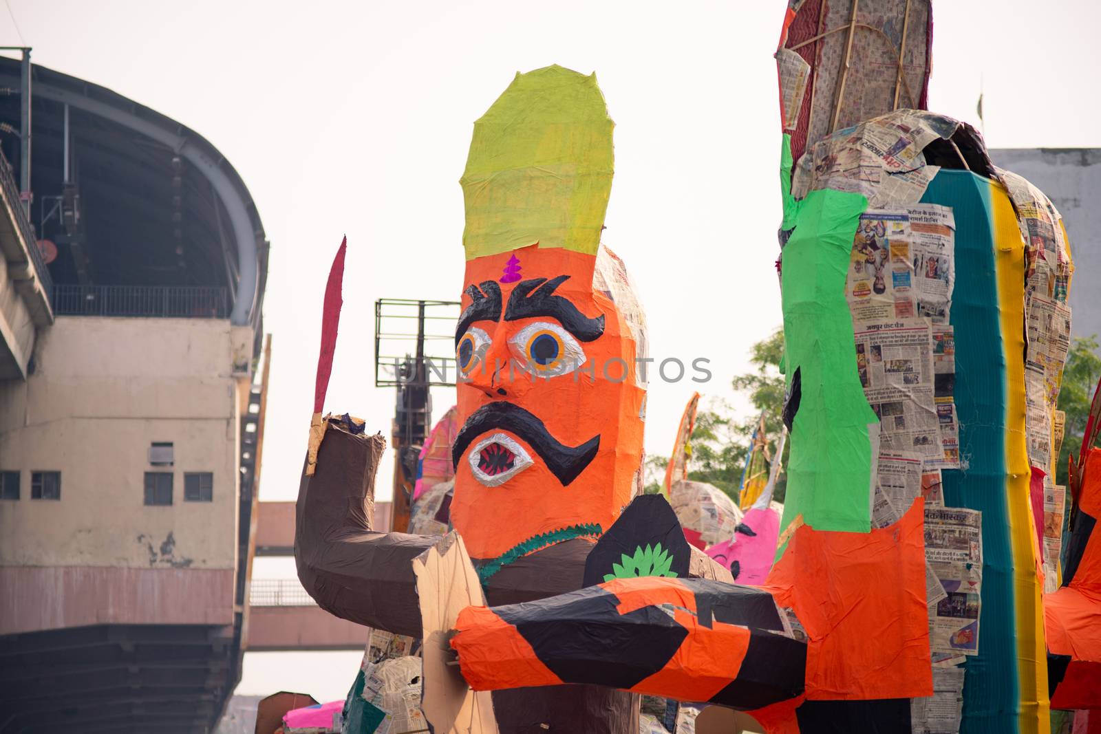 Huge colorful ravana effigy with a sword and a shield placed on the side of a busy street with a tree in the foreground and traffic passing by. Shows the preparations for the hindu festival of dussera prior to diwali celebrating the victory of good over evil as given in the hindu poem epic ramayana