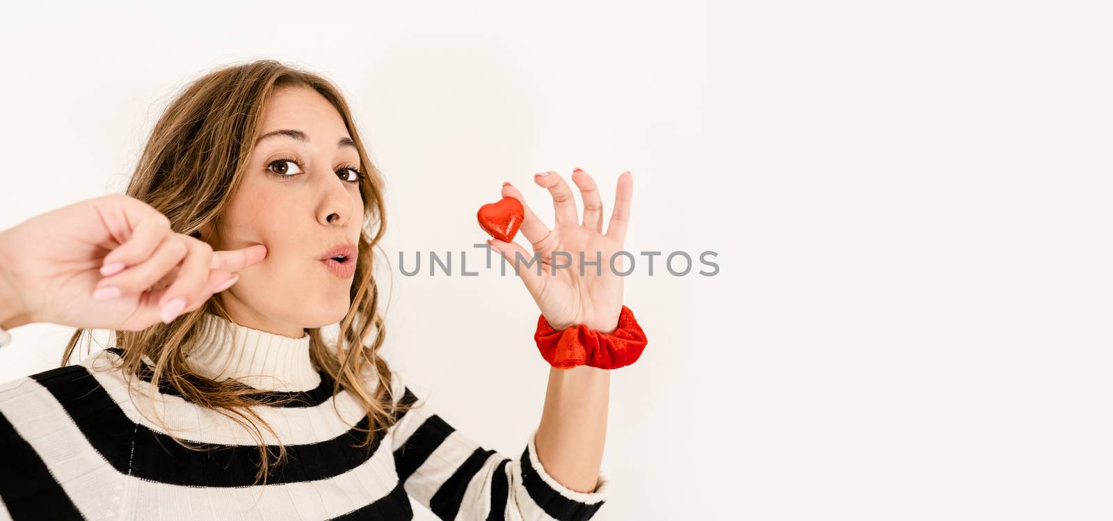 Head and shoulders close up of a blonde cute excited woman looking at the camera holding a heart-shaped chocolate between your fingers and gluttony expression - Chocolate: symbol of sin and temptation by robbyfontanesi