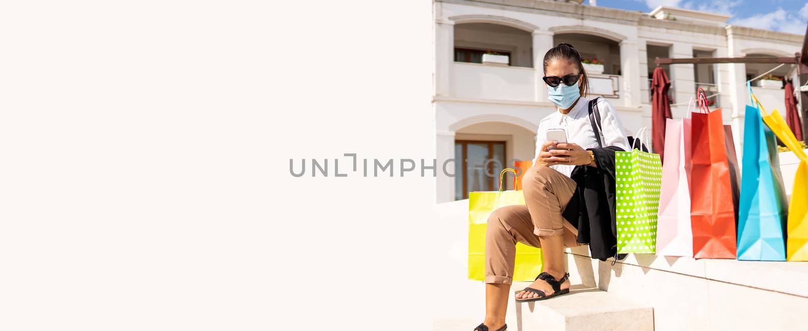 Young cute stylish woman browsing with her smartphone wearing a medical mask sitting among her colored shopping bags - New normal buying activity with remote connection and Coronavirus diseases by robbyfontanesi