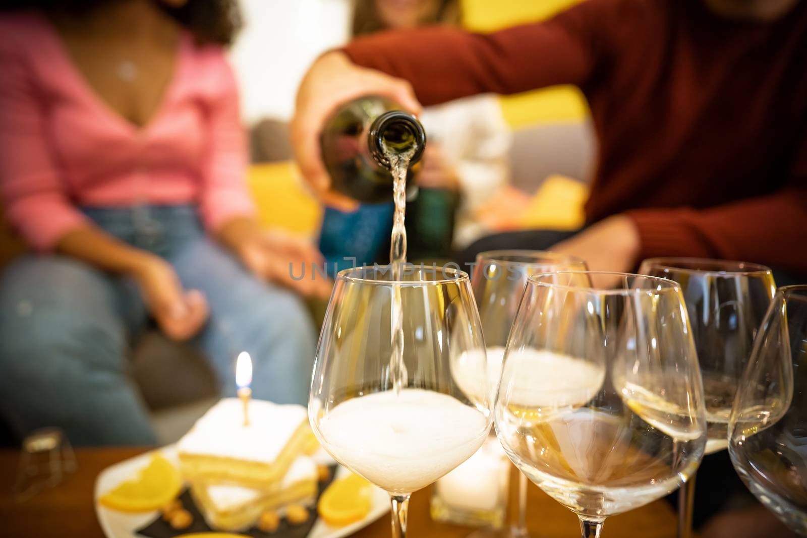 Close up of sparkling white wine being poured in a goblet among other glasses and a birthday cake - Unrecognizable young people celebrating at home drinking champagne for a happy event