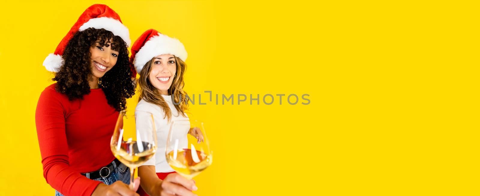 Toasting to Christmas together regardless of diversity - Two women, black Hispanic and Caucasian wearing Santa hat, looking at the camera holding a glass of white wine on big yellow copy space