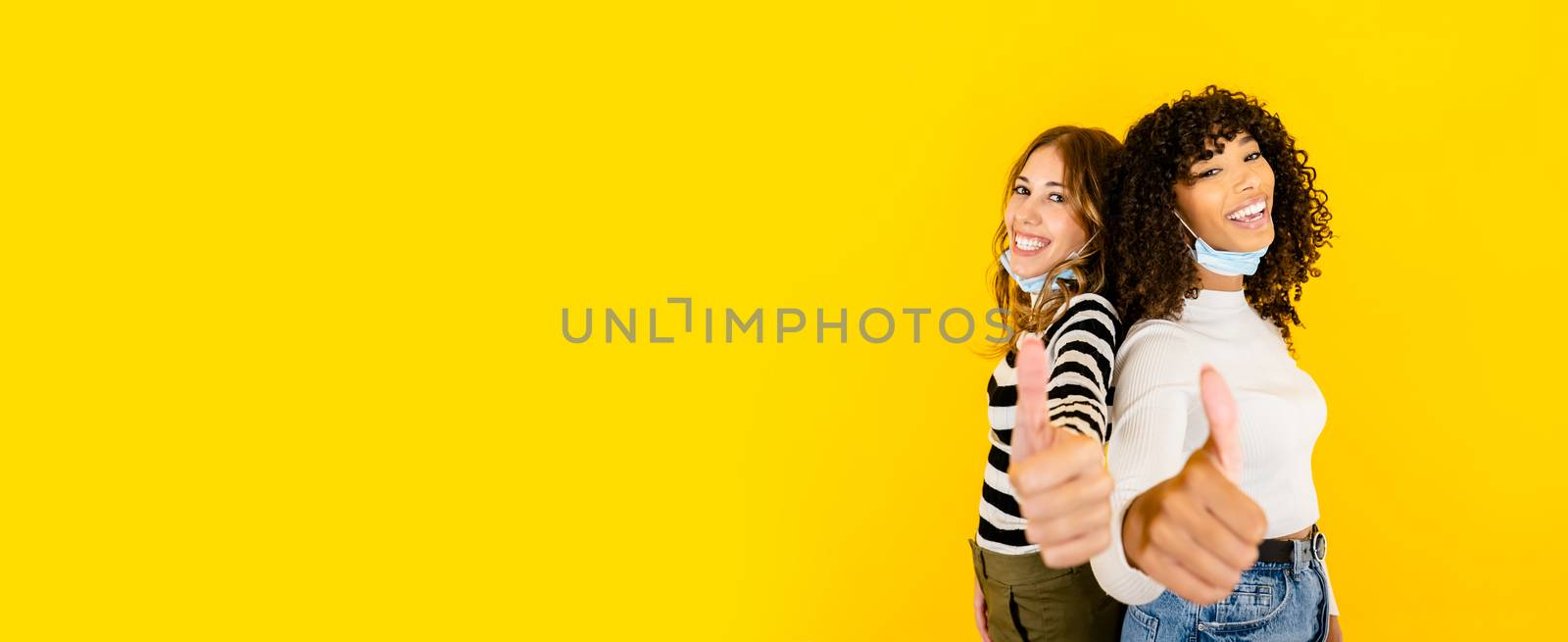 Two women black Hispanic and Caucasian with medical mask down looking at the camera with thumbs up on big yellow copy space - Together we can regardless of troubles - Selective focus on curly brunette by robbyfontanesi