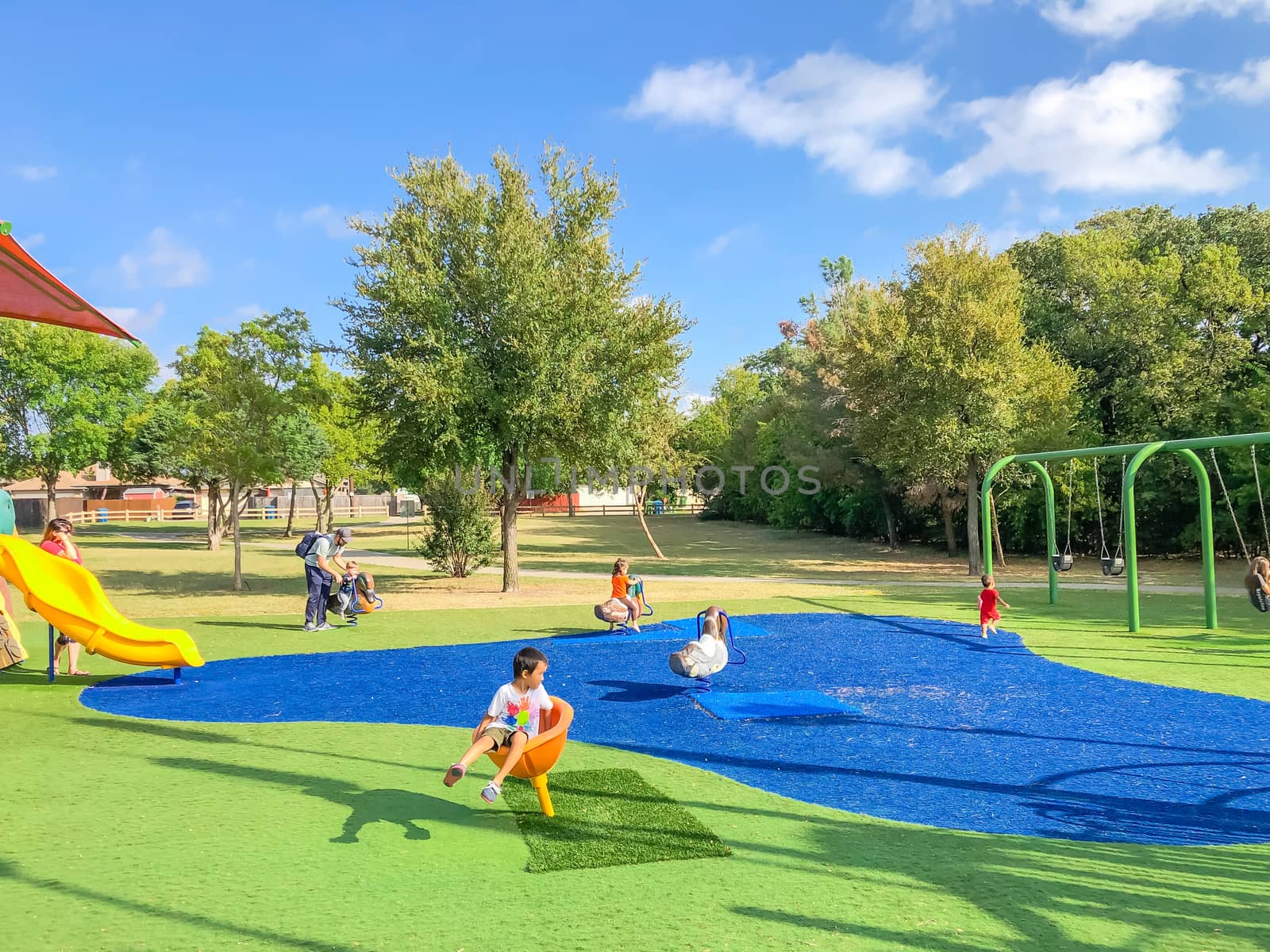 Large neighborhood playground with sun shade sails, artificial grass and kids playing in Flower Mound, Texas, America. Suburban recreational facility surrounded by wooden fence