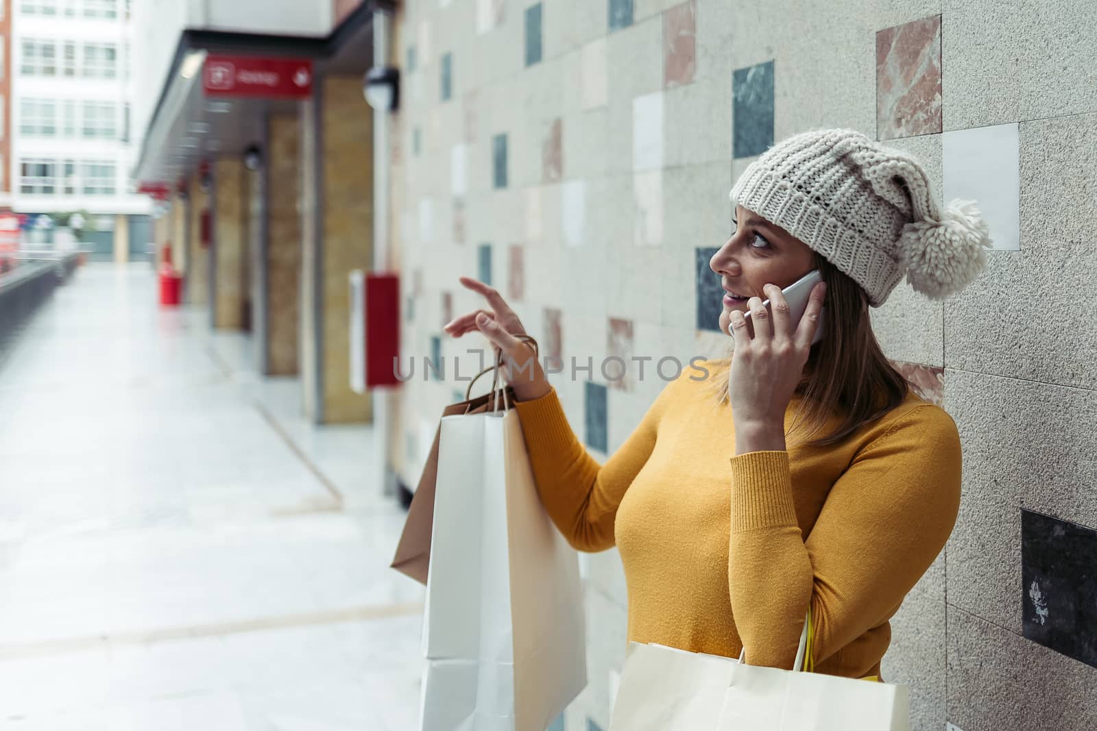 Adult woman wearing a yellow sweater and wool cap talking on her mobile phone while holding shopping bags.