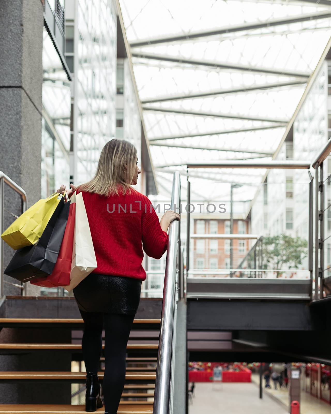 Adult woman wearing a red sweater walking up the stairs of a shopping mall carrying colorful shopping bags.