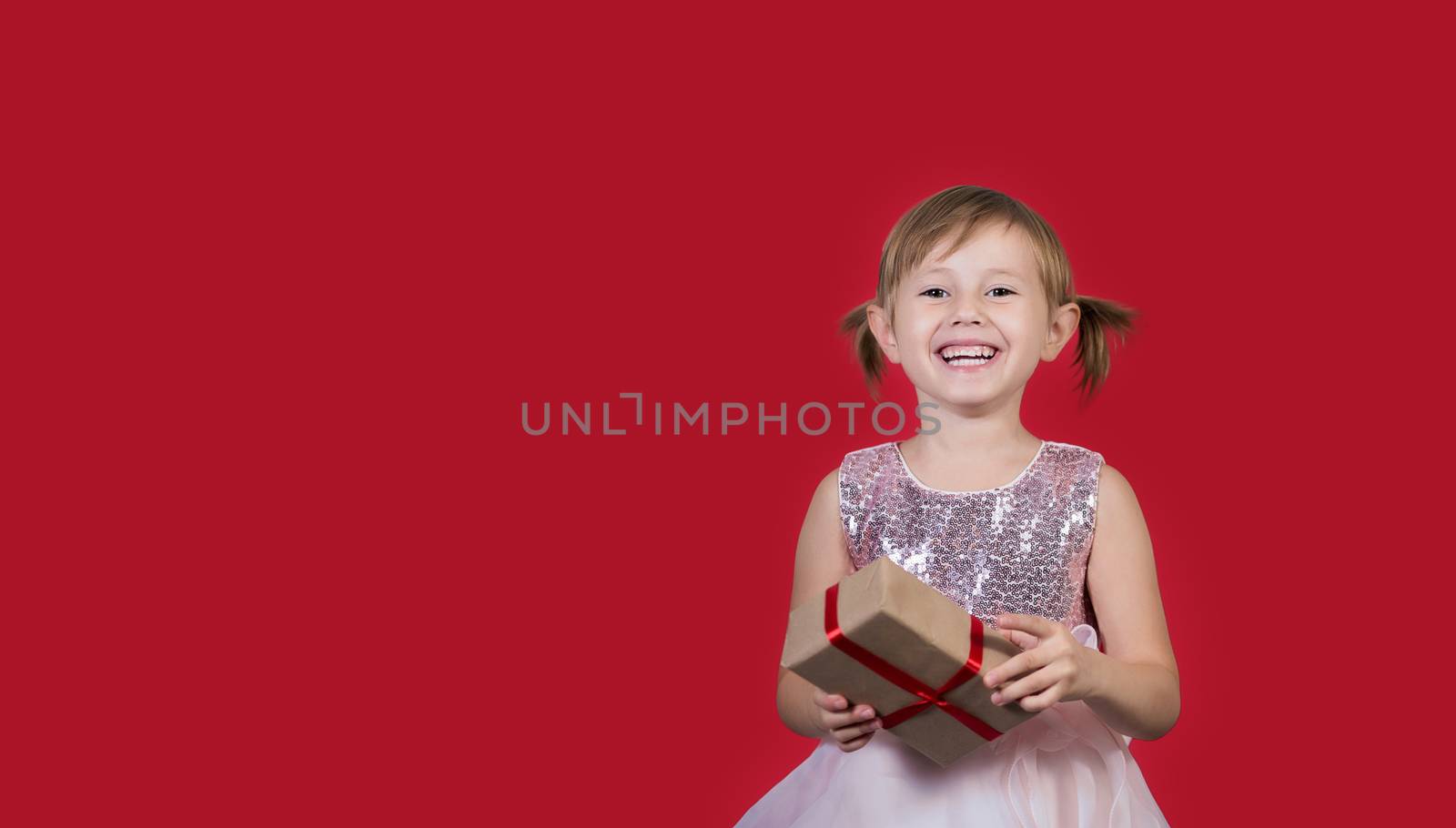 Smiling cute cacusian little girl in santa hat looking at the camera with gift box celebrating happy 2021 New Year isolated on red background. Merry Christmas presents shopping sale.