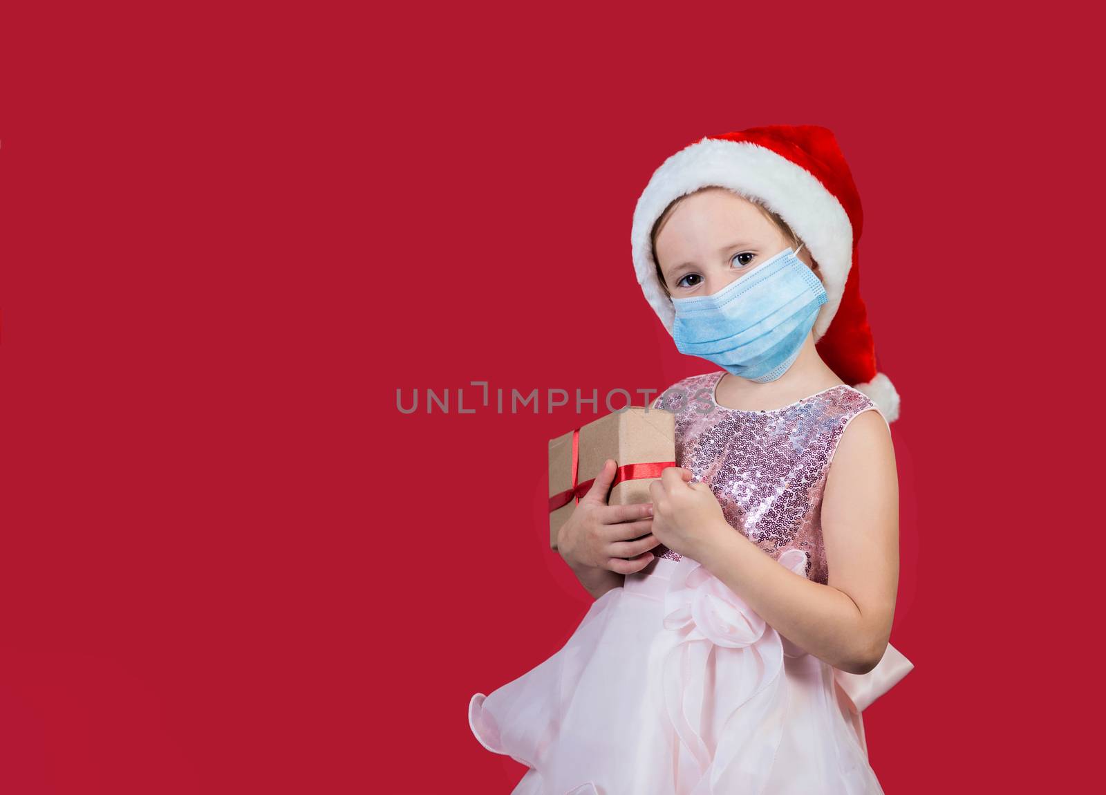 Adorable cacusian little girl wearing medical mask in santa hat with gift box celebrating happy New Year isolated on red background. Merry Christmas presents shopping sale.