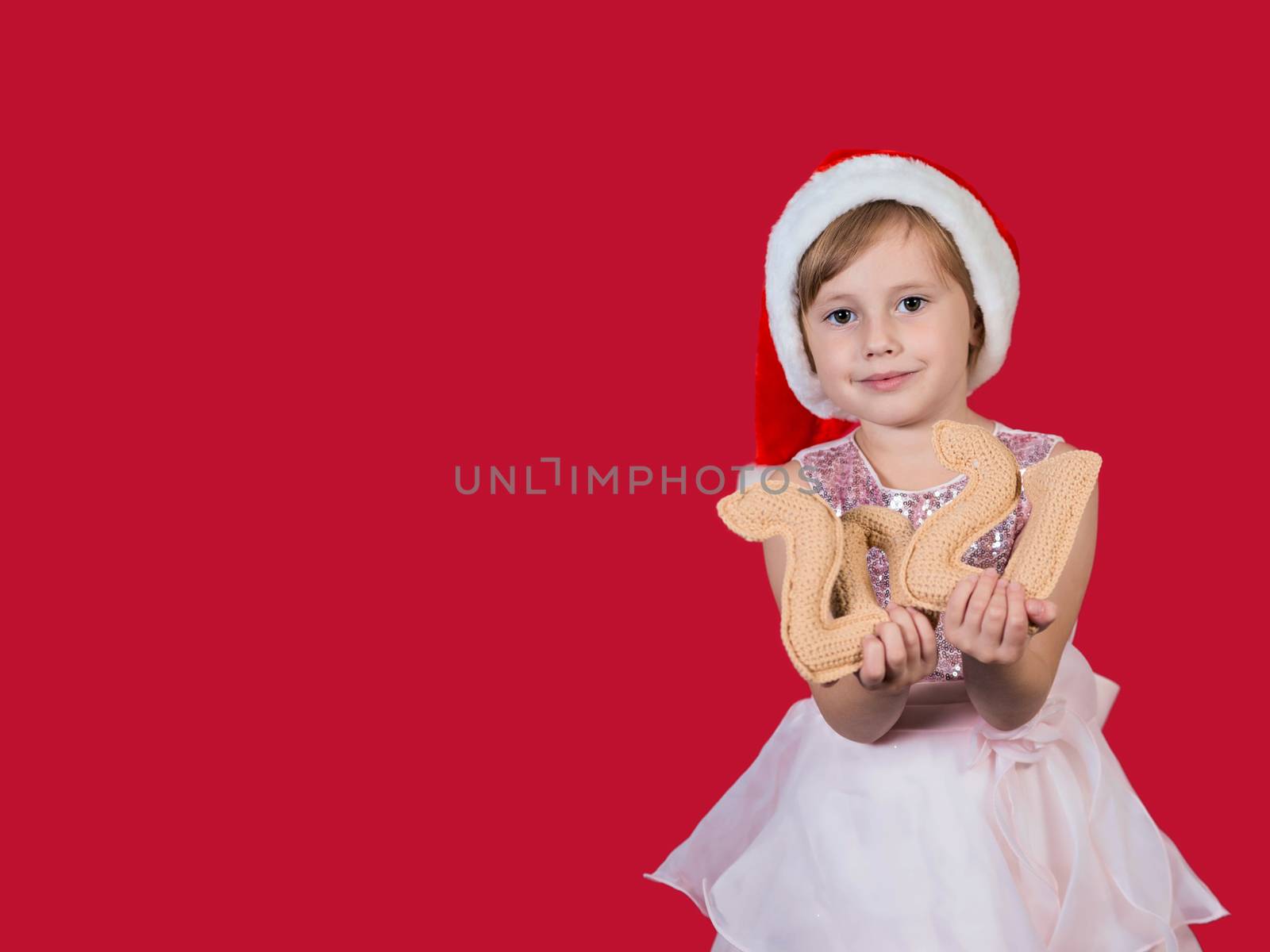 Adorable funny cacusian little girl in santa hat looking at the camera with croched numbers 2021 celebrating happy New Year isolated on red background. Merry Christmas presents shopping sale.