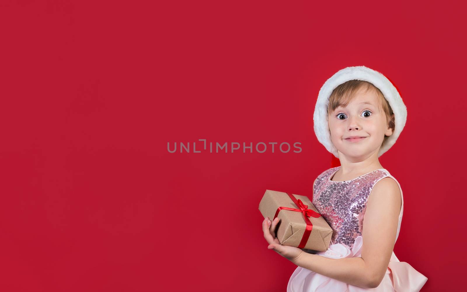 Surprised funny cacusian cute child girl in santa hat looking at the camera with gift box celebrating happy 2021 New Year isolated on red background. Merry Christmas presents shopping sale.