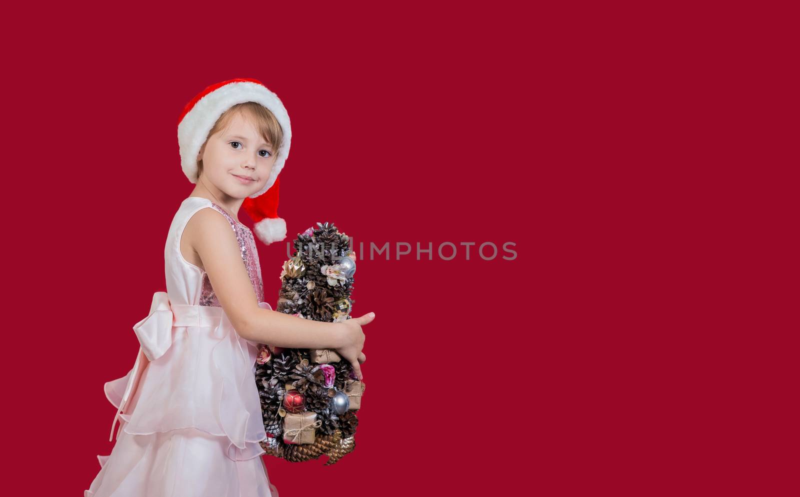 Beautiful cute cacusian little girl in santa hat looking at the camera with homemade christmas tree celebrating happy 2021 New Year isolated on red background. Merry Christmas presents shopping sale.