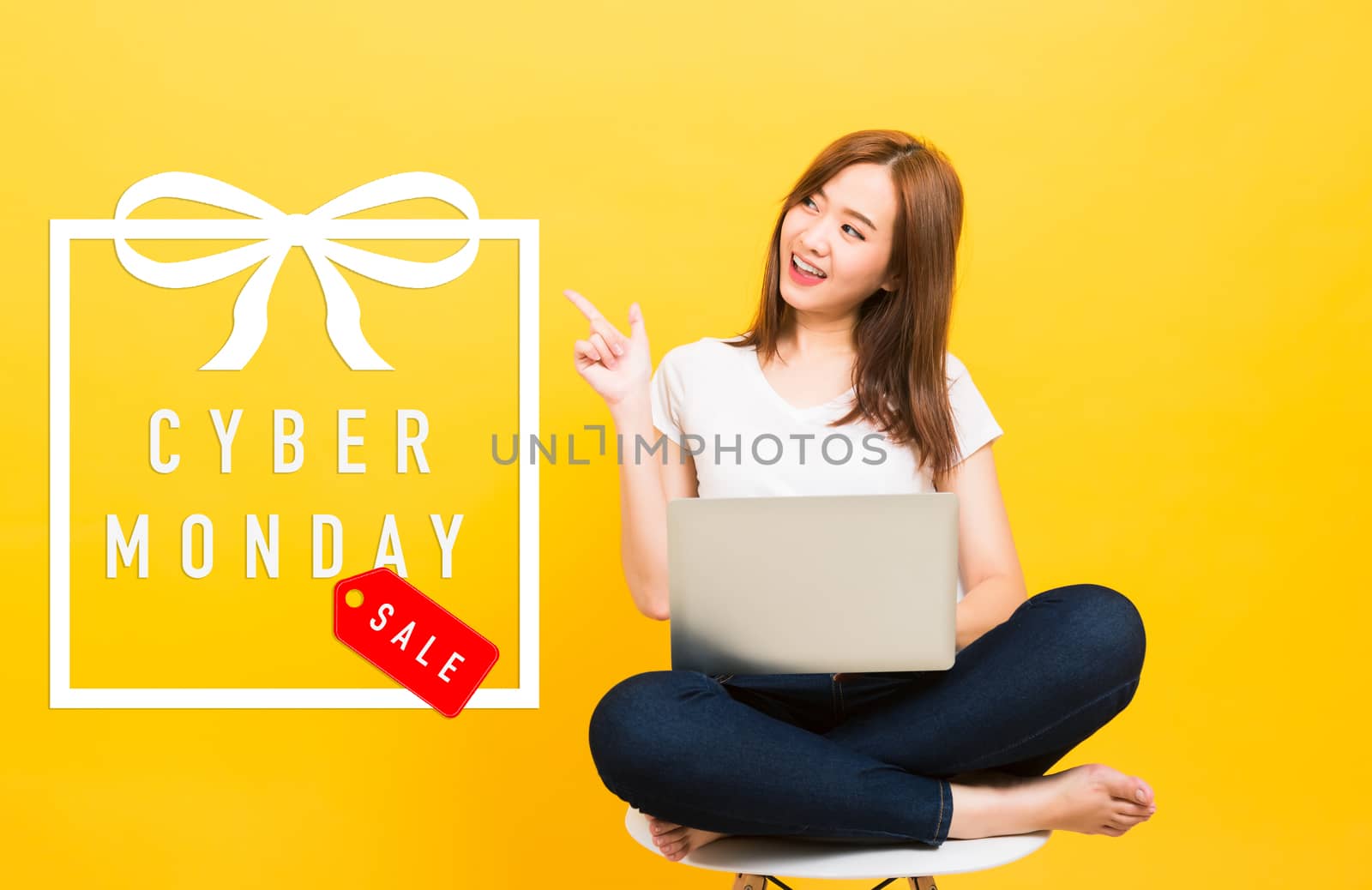 Asian happy beautiful young woman smiling sitting crossed legs on chair use laptop computer point finger to Cyber Monday text in gift box looking to side isolated, studio shot yellow background