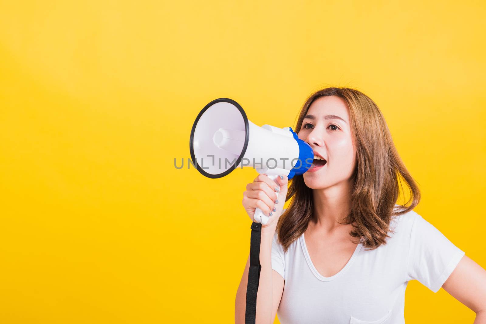 Asian Thai happy portrait beautiful cute young woman stand to make announcement message shouting screaming in megaphone looking to side away, studio shot isolated on yellow background with copy space