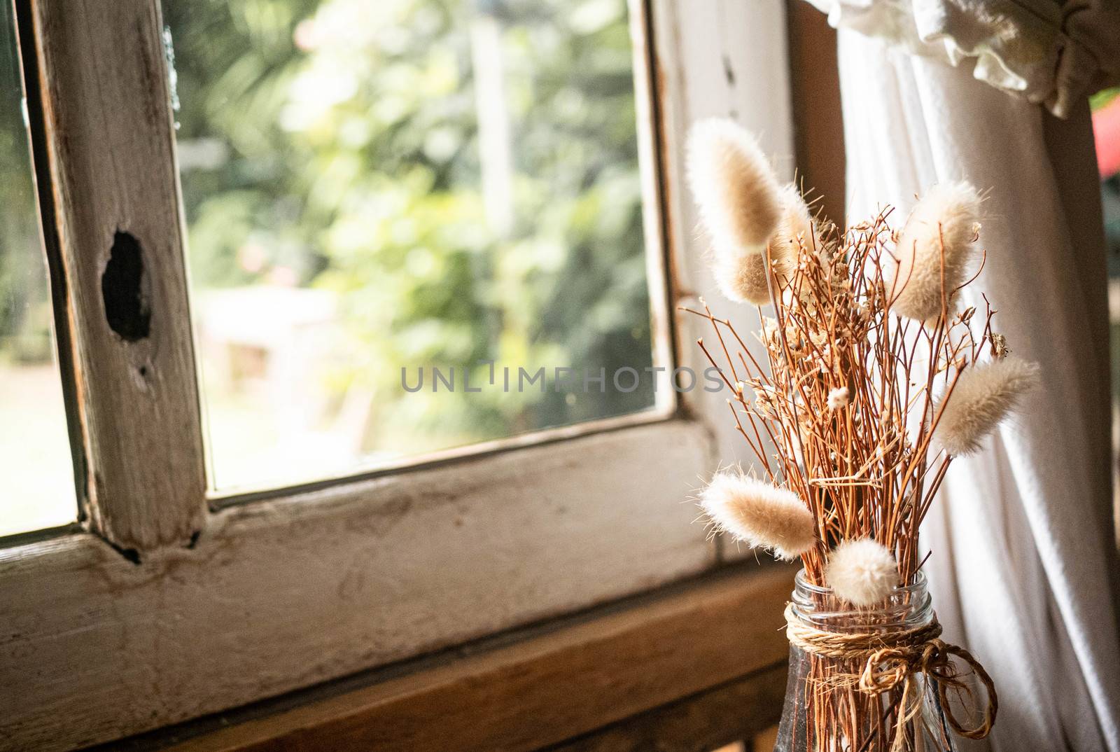 Dried flowers in a glass bottle on wooden table by the window wi by TEERASAK