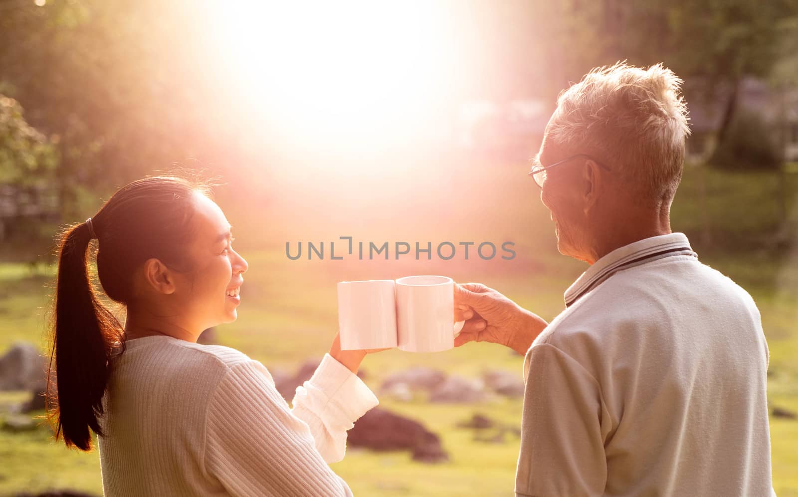 Rear view of granddaughter and grandfather drinking hot coffee outdoor and enjoying landscape during holiday on the mountain with sunlight in morning.