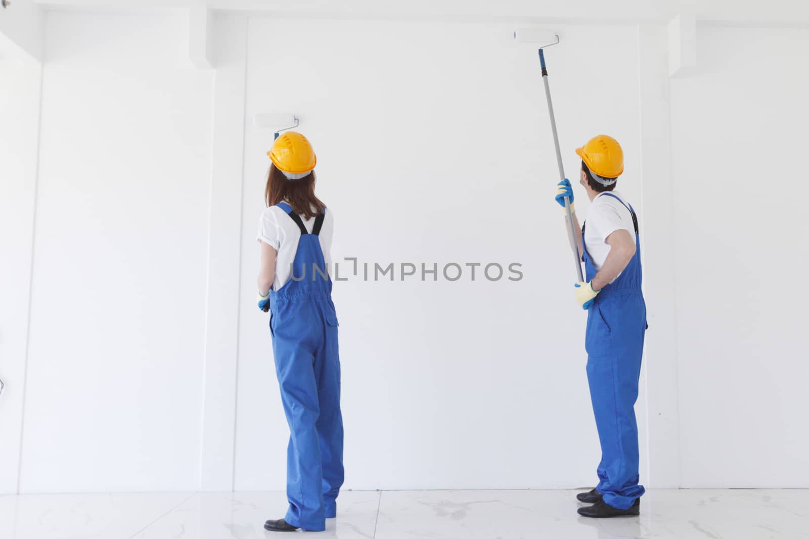 Two young workers in uniform of coverall and hardhat painting white wall