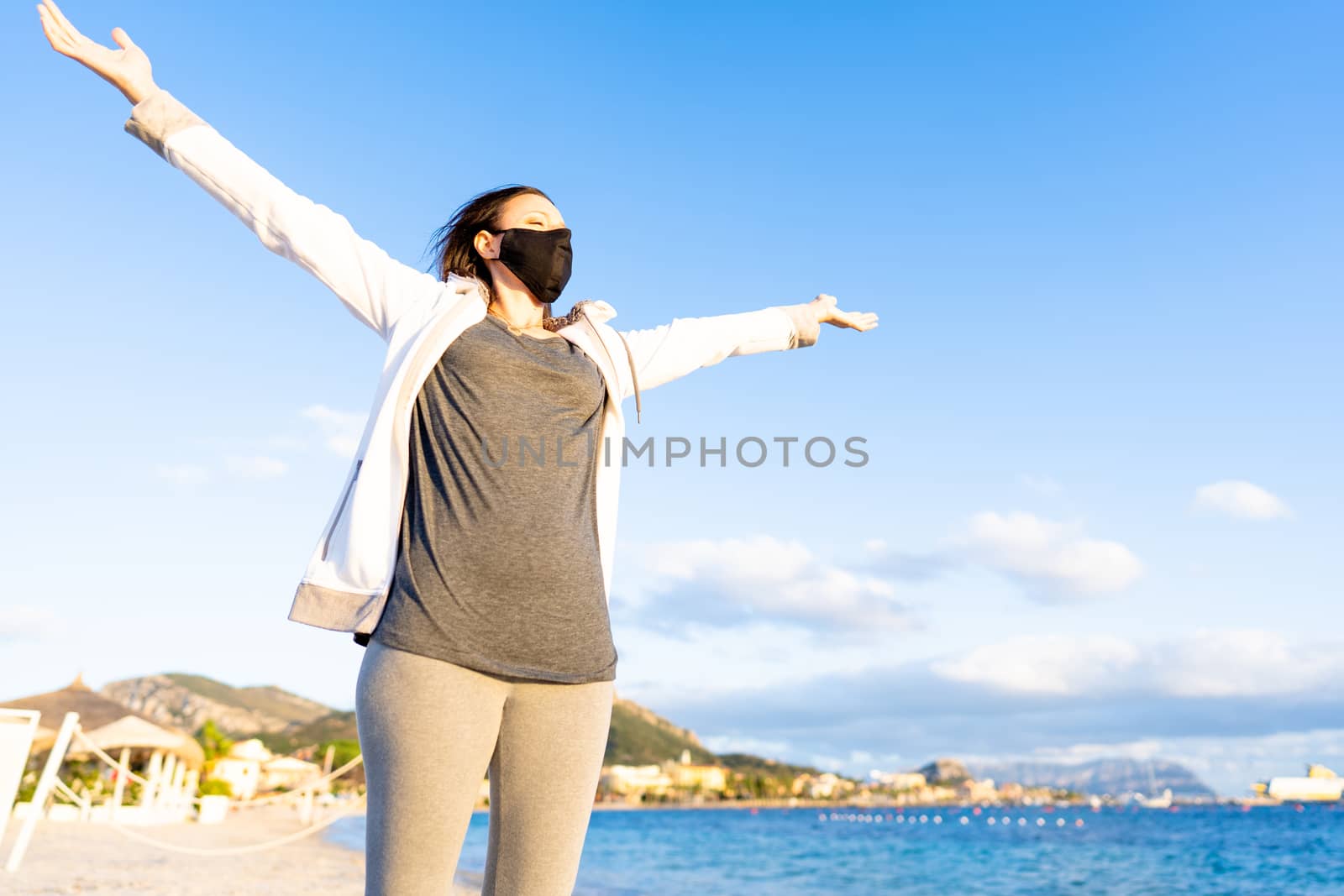 Young woman wearing black medical mask spreading her arms to the blue sky looking to the setting sun in Golfo Aranci, Sardinia - Pleasant lifestyle at the sea in winter despite the Coronavirus