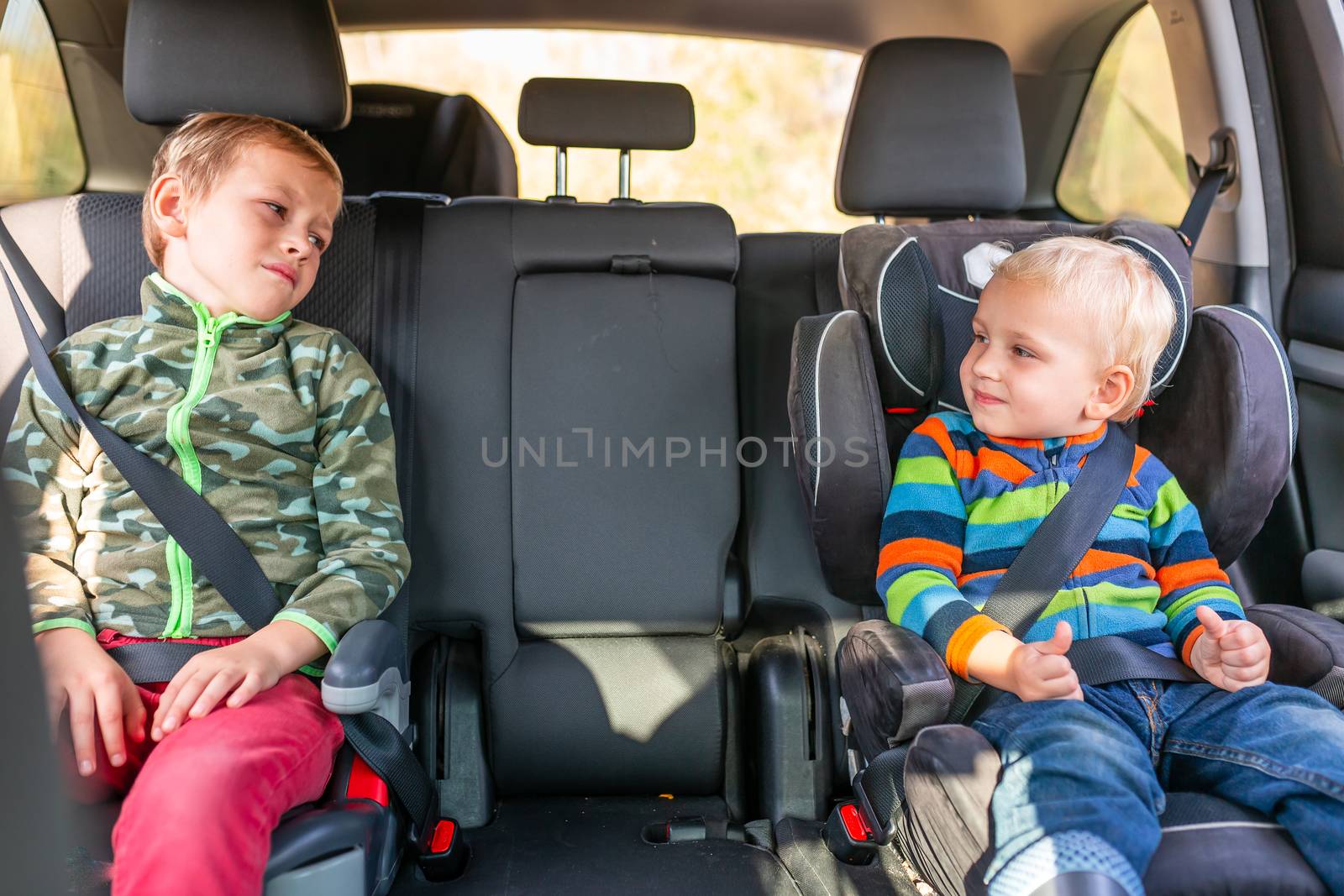 Two little boys sitting on a car seat and a booster seat buckled up in the car. Children's Car Seat Safety