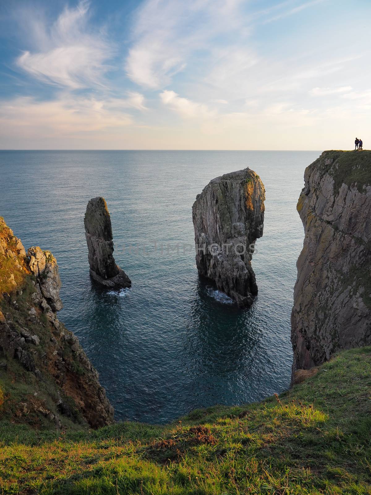 Elegug Stacks, two prominent limestone sea stacks rise up from the sea close to the cliffs, with two people looking down over them, south coast of Pembrokeshire, Wales, UK
