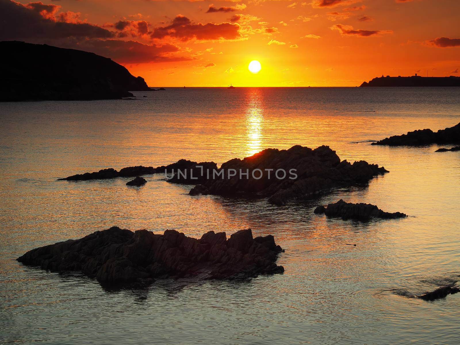 Stunning orange sunset looking out over rocks to West Angle Bay with fishing boat passing by in the background, south Pembrokeshire coast, Wales, UK